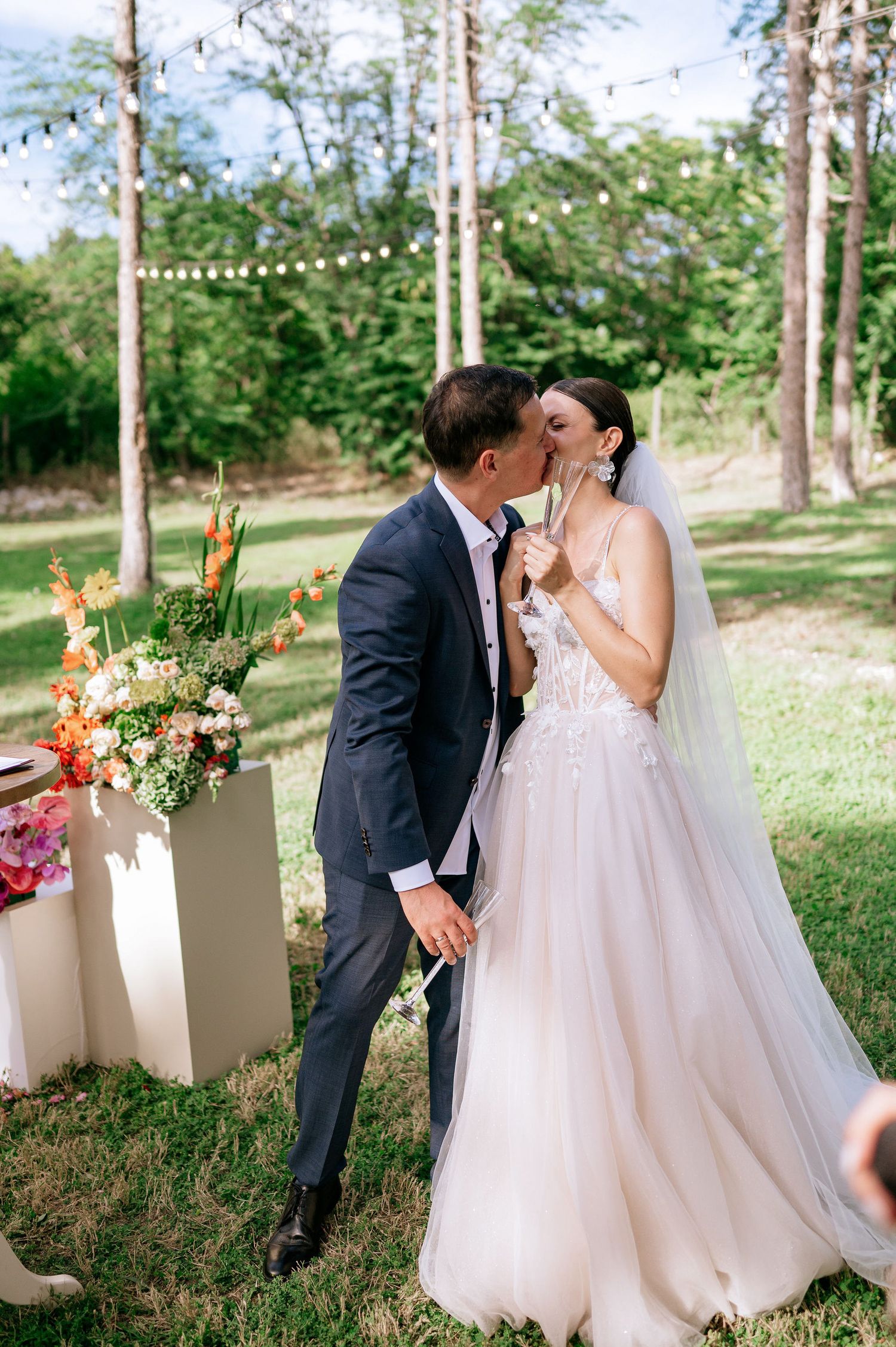 Romantic outdoor wedding scene with couple embracing among tall trees and floral arrangements.