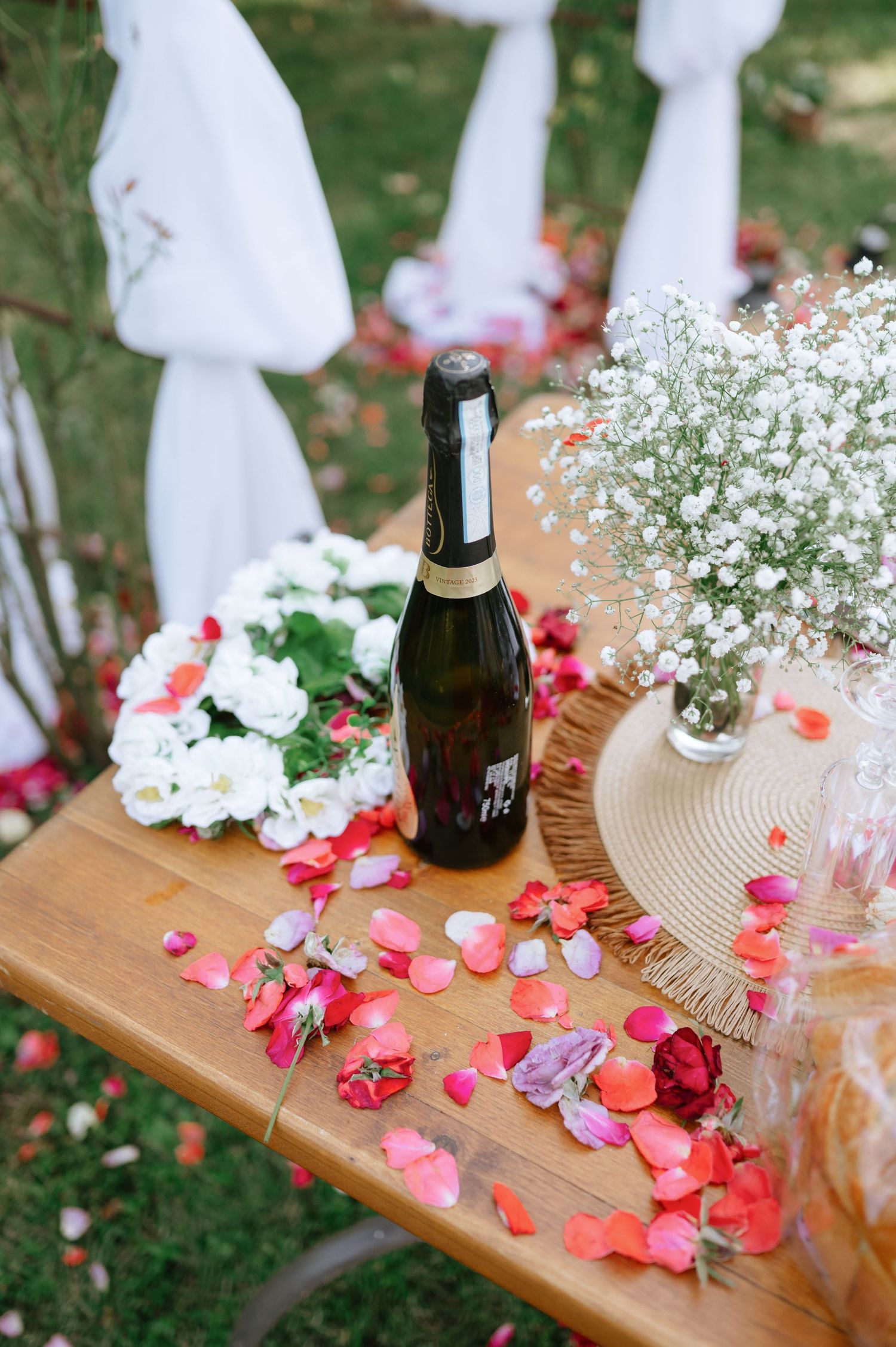 Wedding reception table decorated with rose petals, baby's breath flowers and wine bottles on wooden surface.