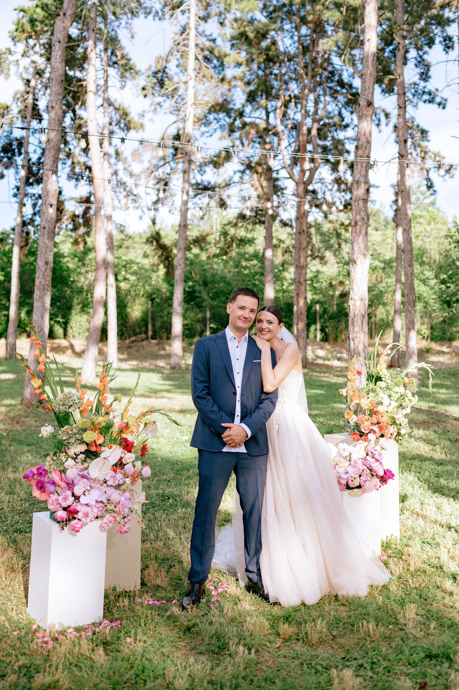 A wedding portrait in a forest setting with pink and orange floral arrangements framing the scene.