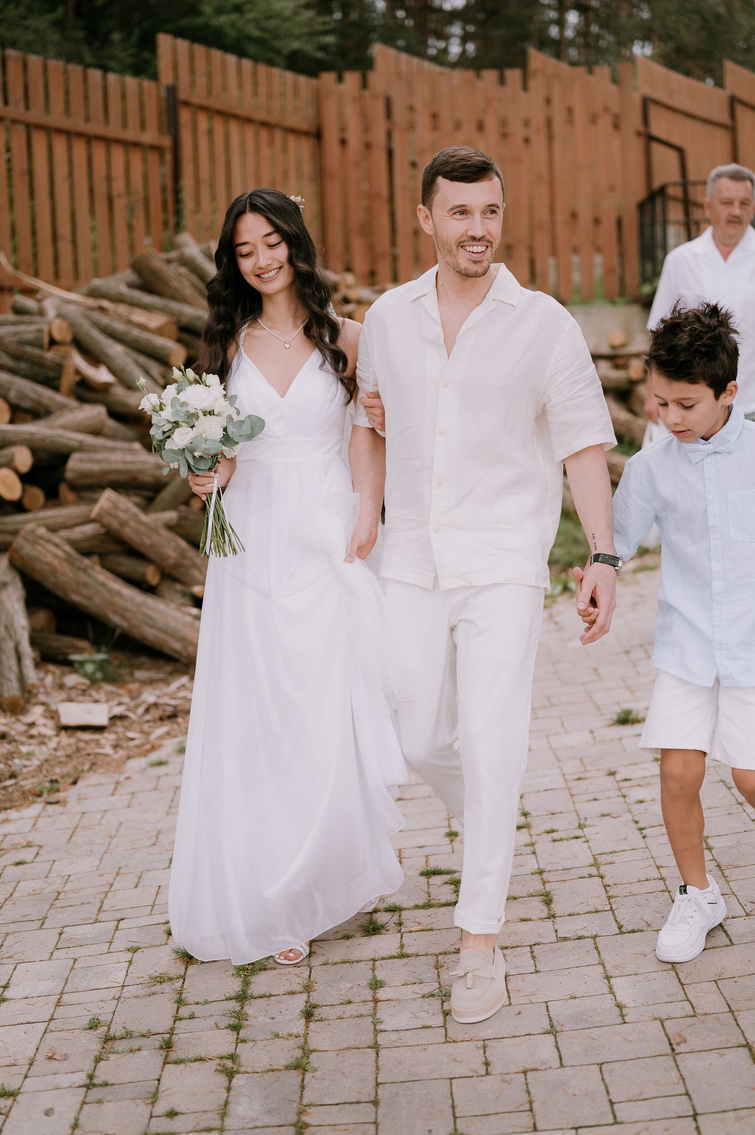 Wedding party dressed in white walking along stone pathway near wooden fence.