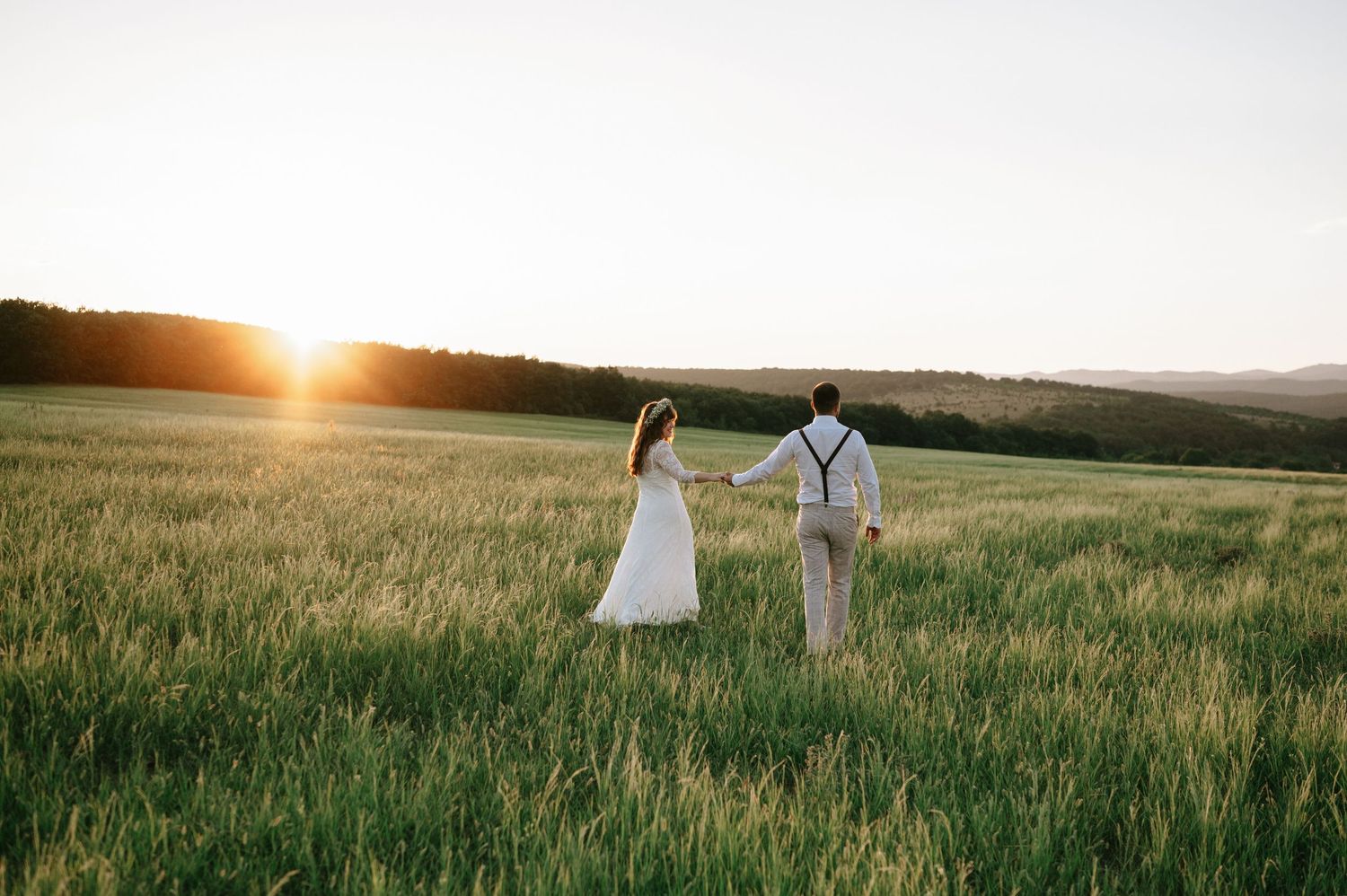 Couple holding hands in a sunlit meadow during golden hour with mountains in the background.