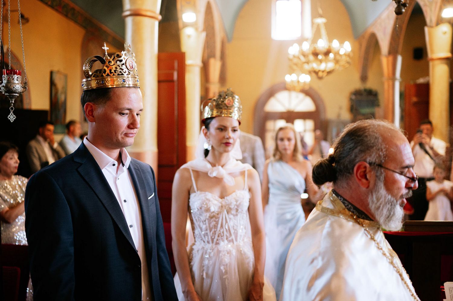 Wedding ceremony participants gathered in an ornate church with chandeliers and painted walls.