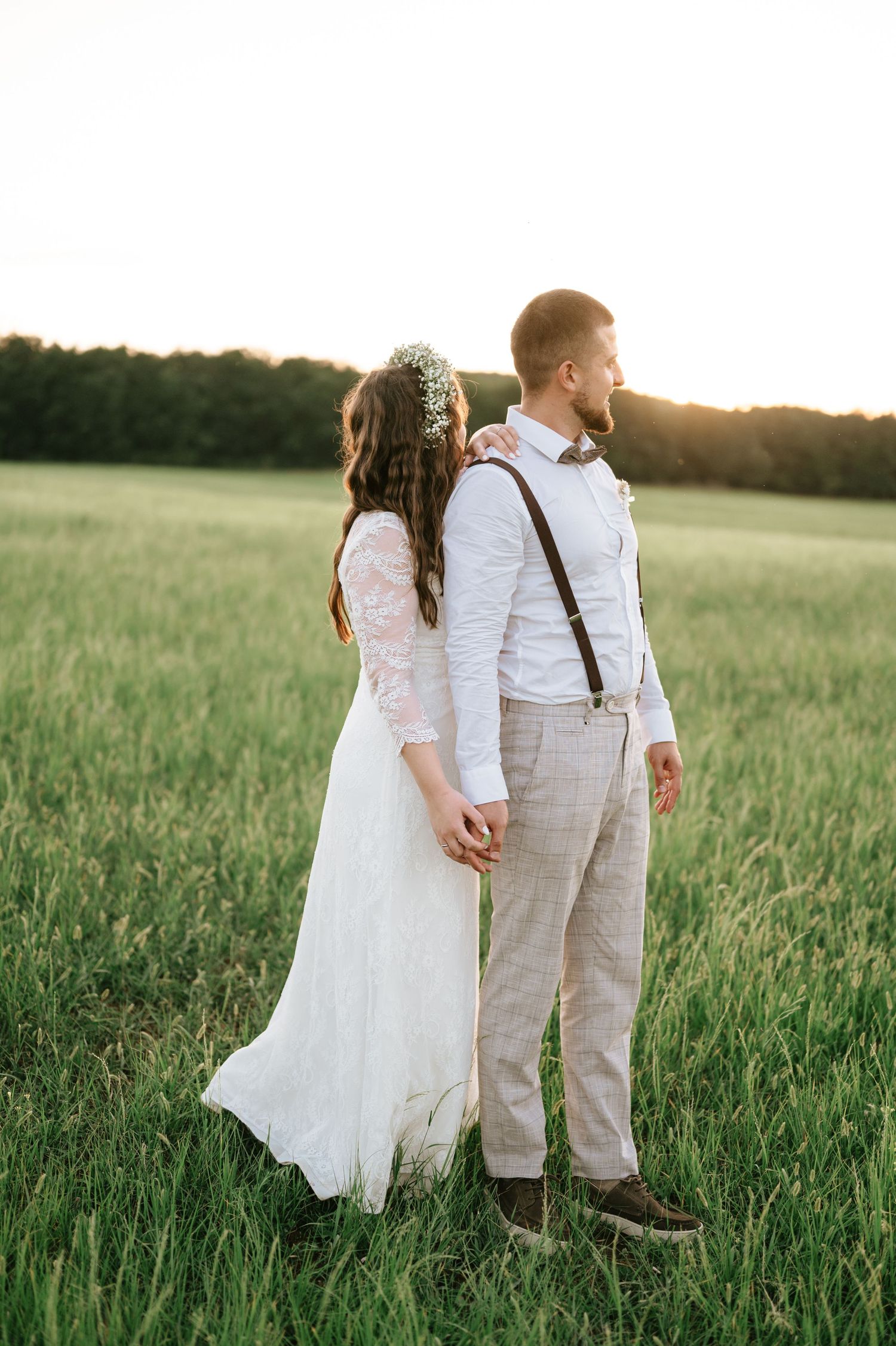 A couple in wedding attire stands in a golden meadow at sunset with mountains in the background.