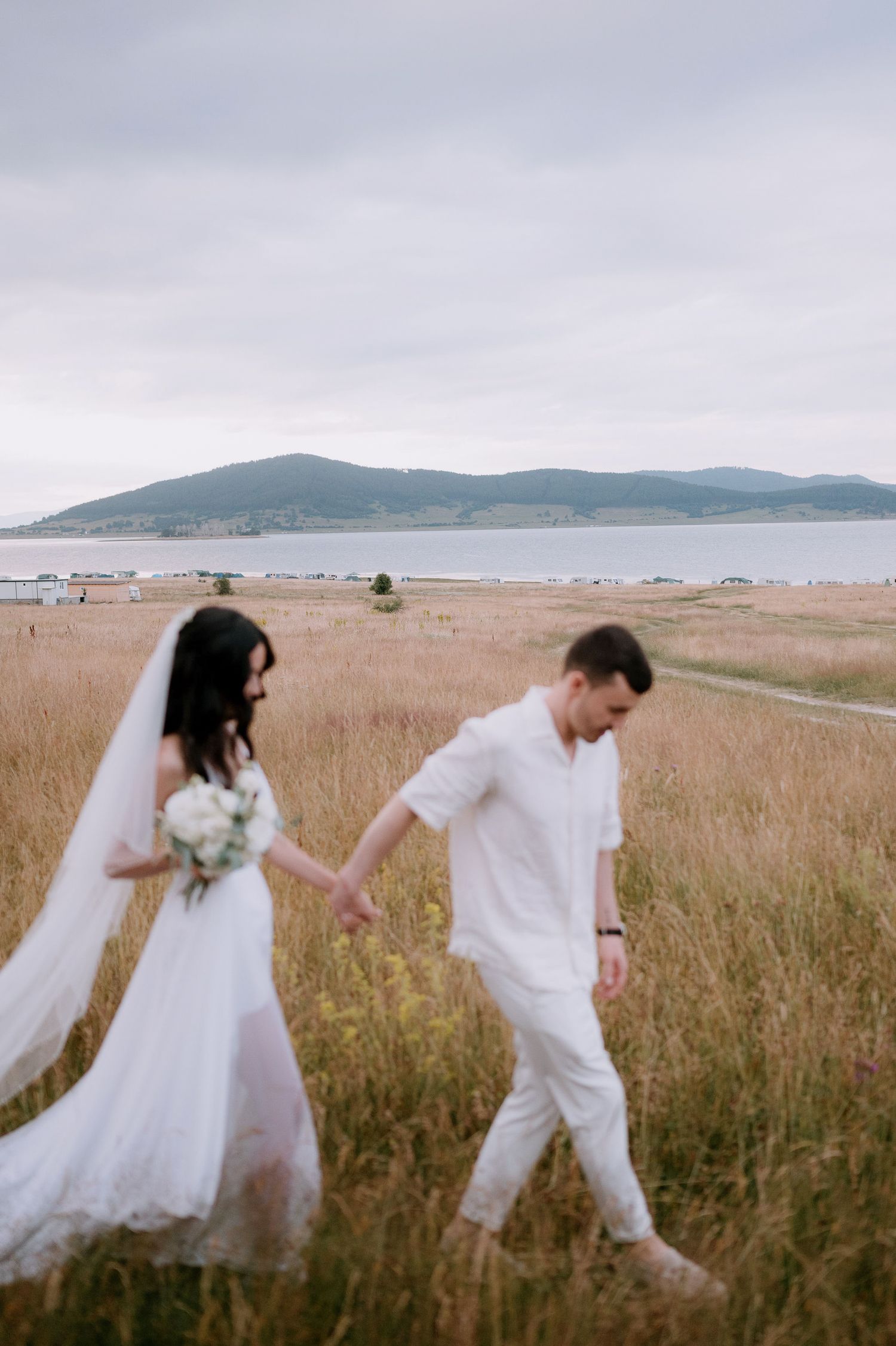 Couple strolling through seaside grassland with mountains visible in distance.