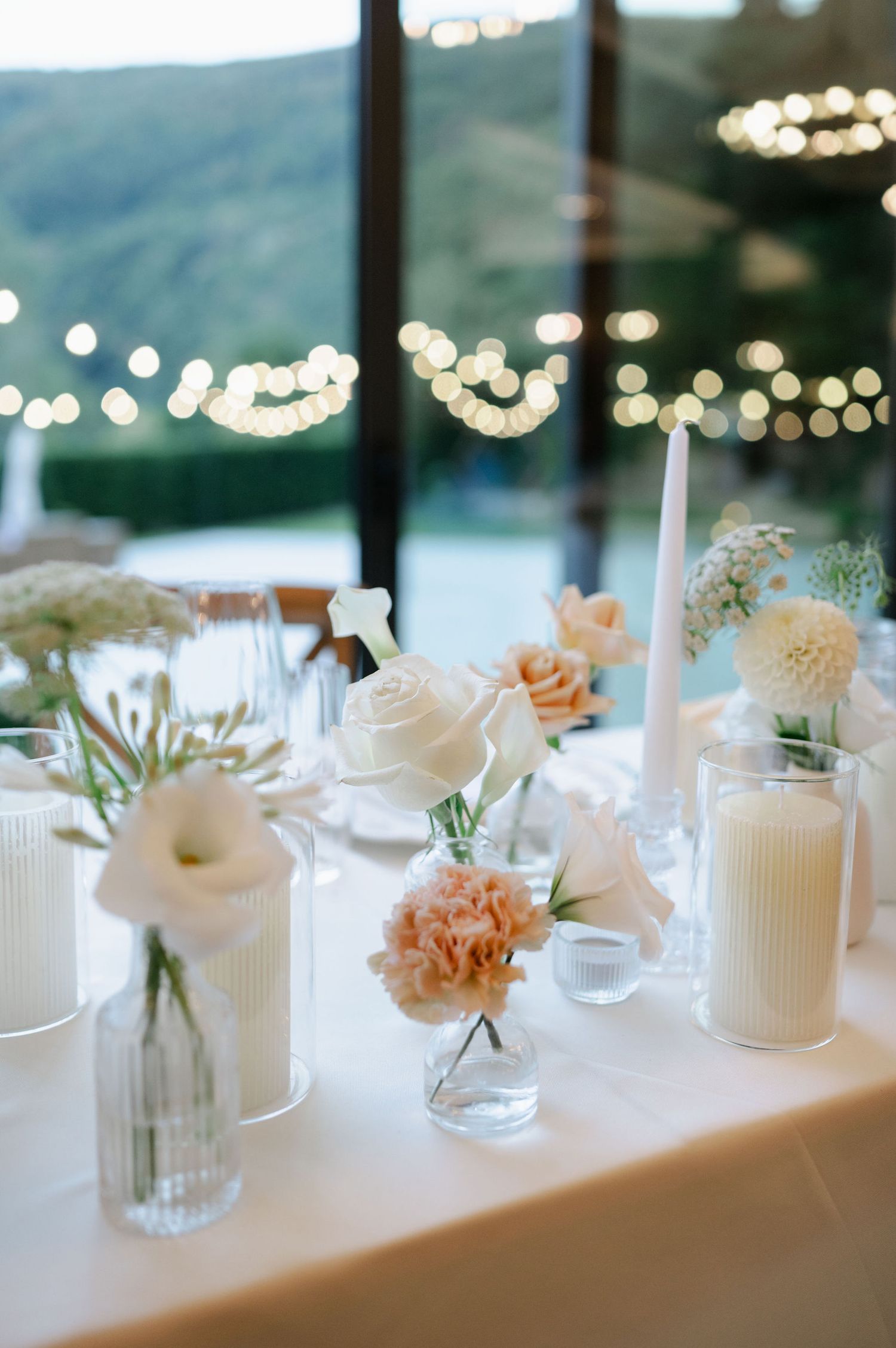 Romantic table settings with white candles and peach roses in glass vases against mountain backdrop.