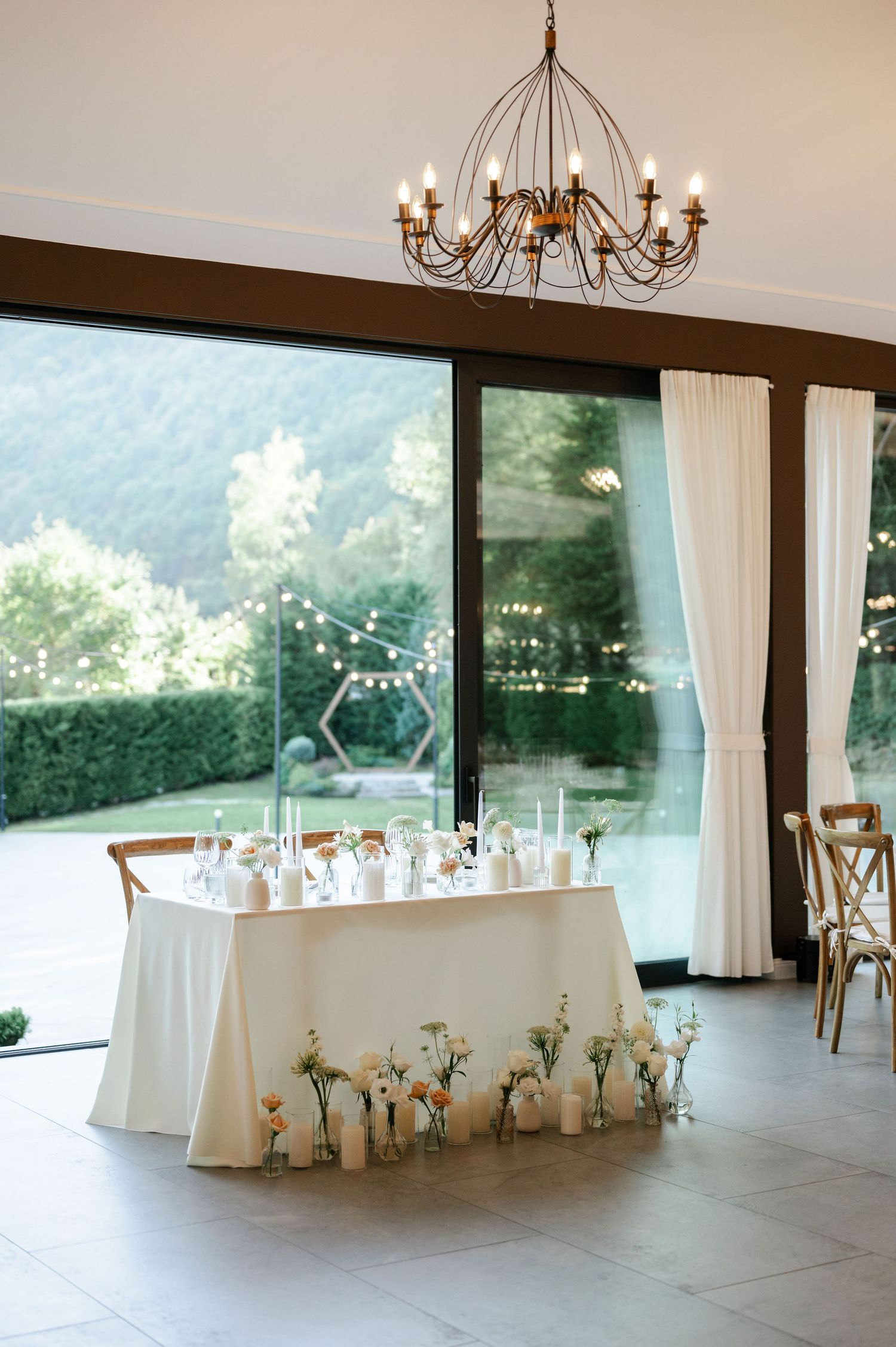 Elegant wedding reception venue with white tablecloths, wooden chairs and large windows overlooking greenery.