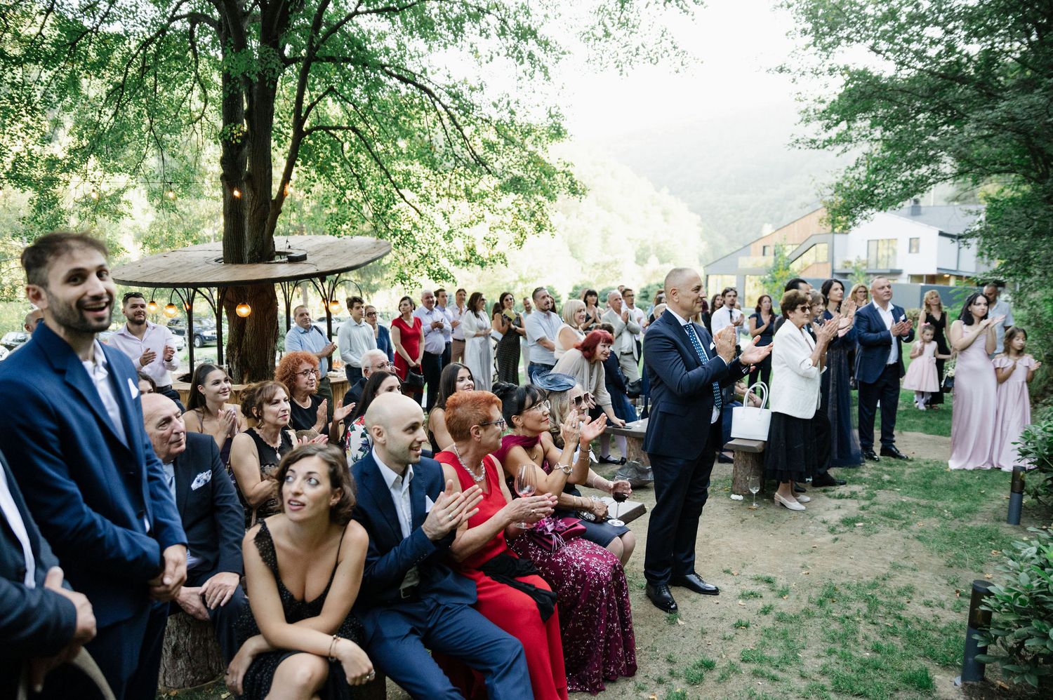 Wedding guests seated outdoors watch ceremony in wooded setting.