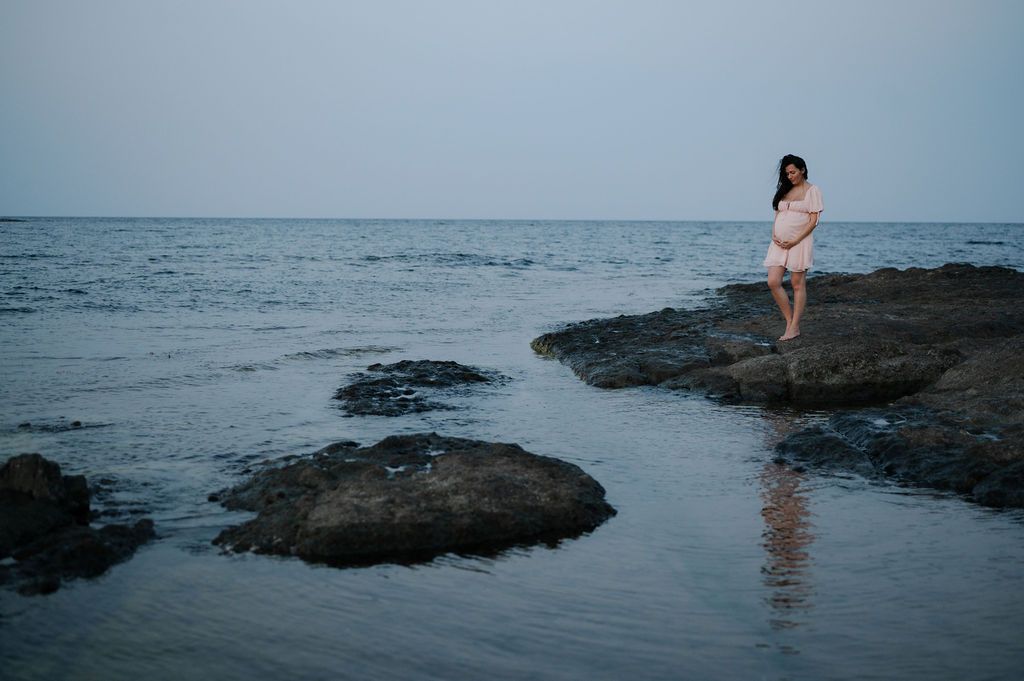 A person stands on dark volcanic rocks at the ocean's edge during sunset creating a serene coastal scene.