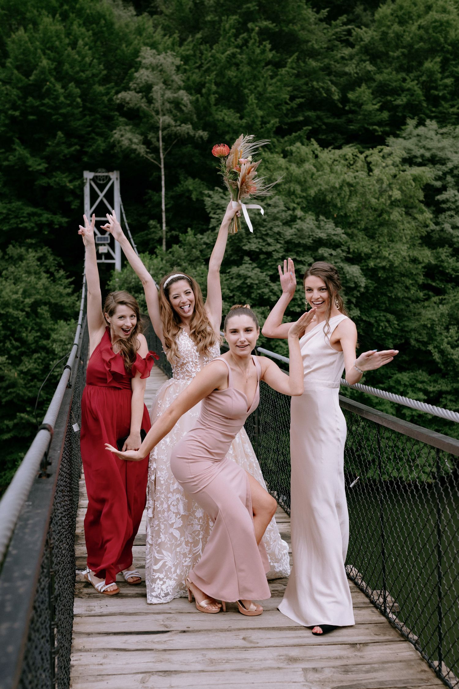 Group in elegant dresses pose together on a wooden suspension bridge surrounded by lush green forest.