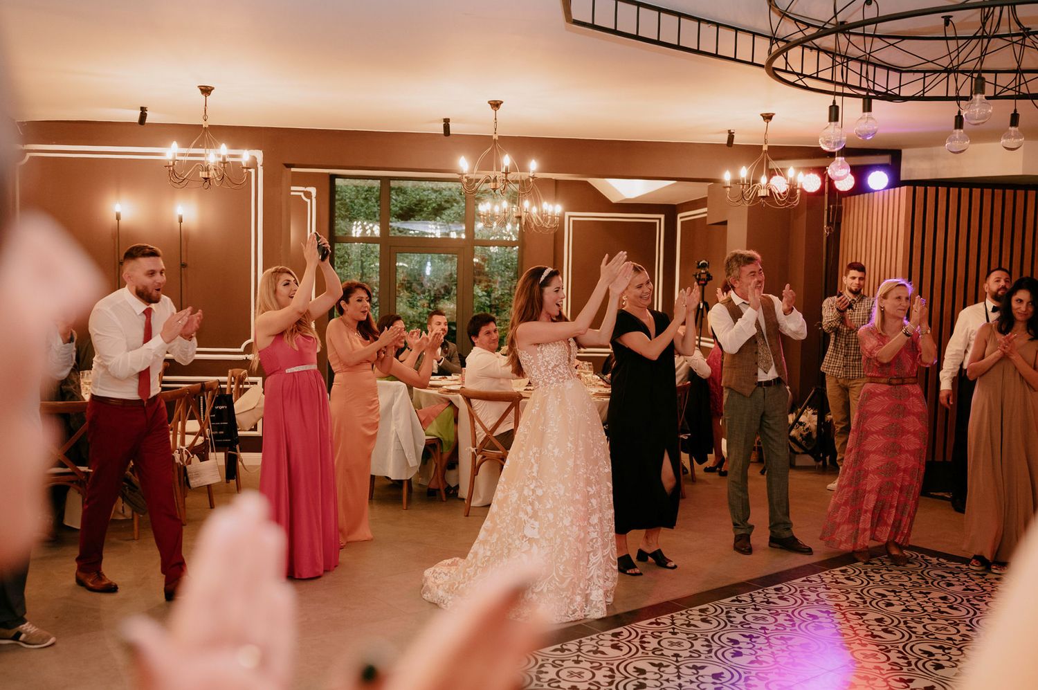 Wedding reception venue features modern chandeliers and wood paneling with guests dancing in a spacious ballroom.