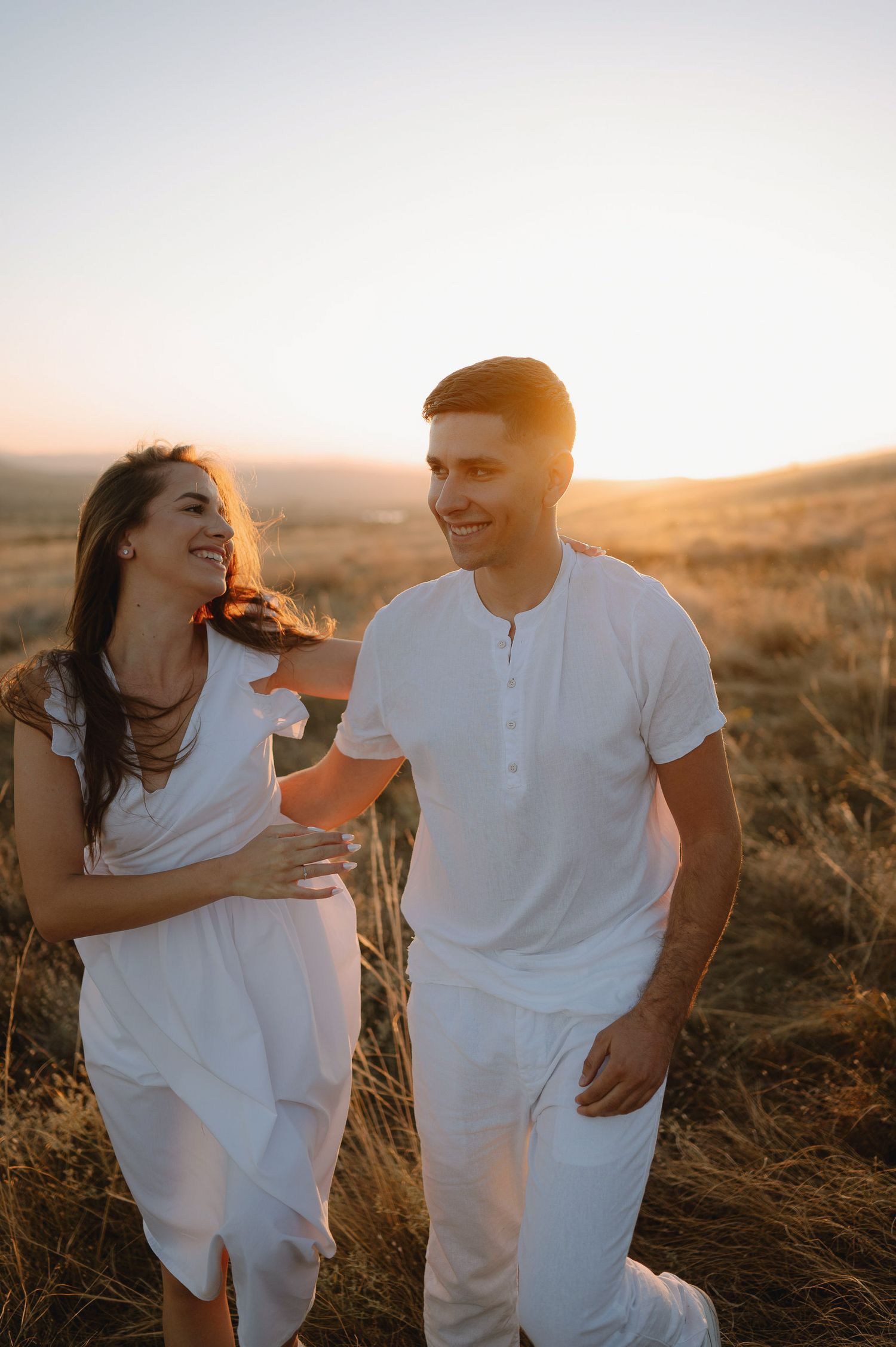 A couple in white outfits laughs together in a golden field at sunset, sharing a romantic moment.