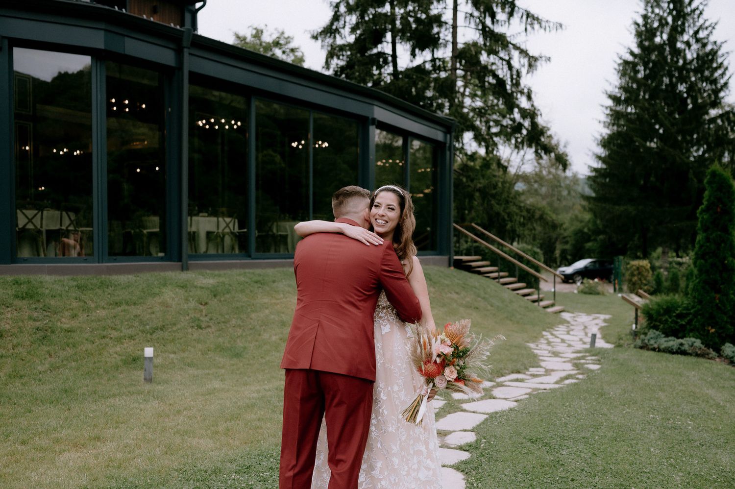 A couple in burgundy wedding attire walks together through a scenic outdoor venue surrounded by evergreen trees.
