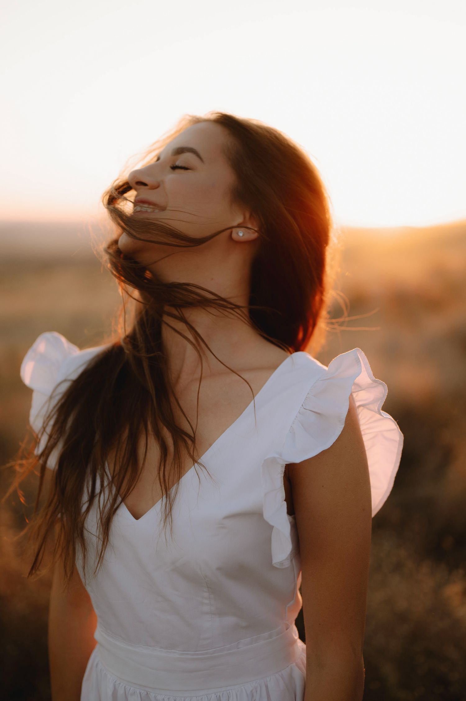 A person in a white ruffled top tilts their head back dramatically in warm golden sunset lighting.