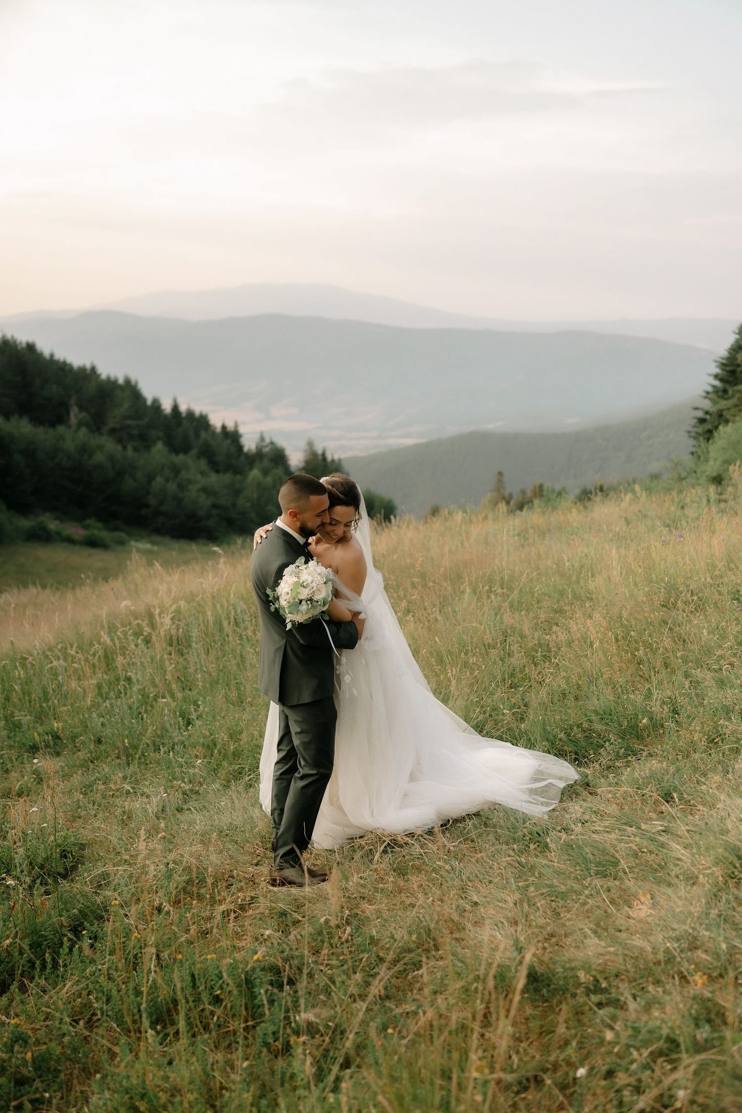 A couple shares an intimate moment in their wedding attire on a grassy hillside with mountain views in the distance.