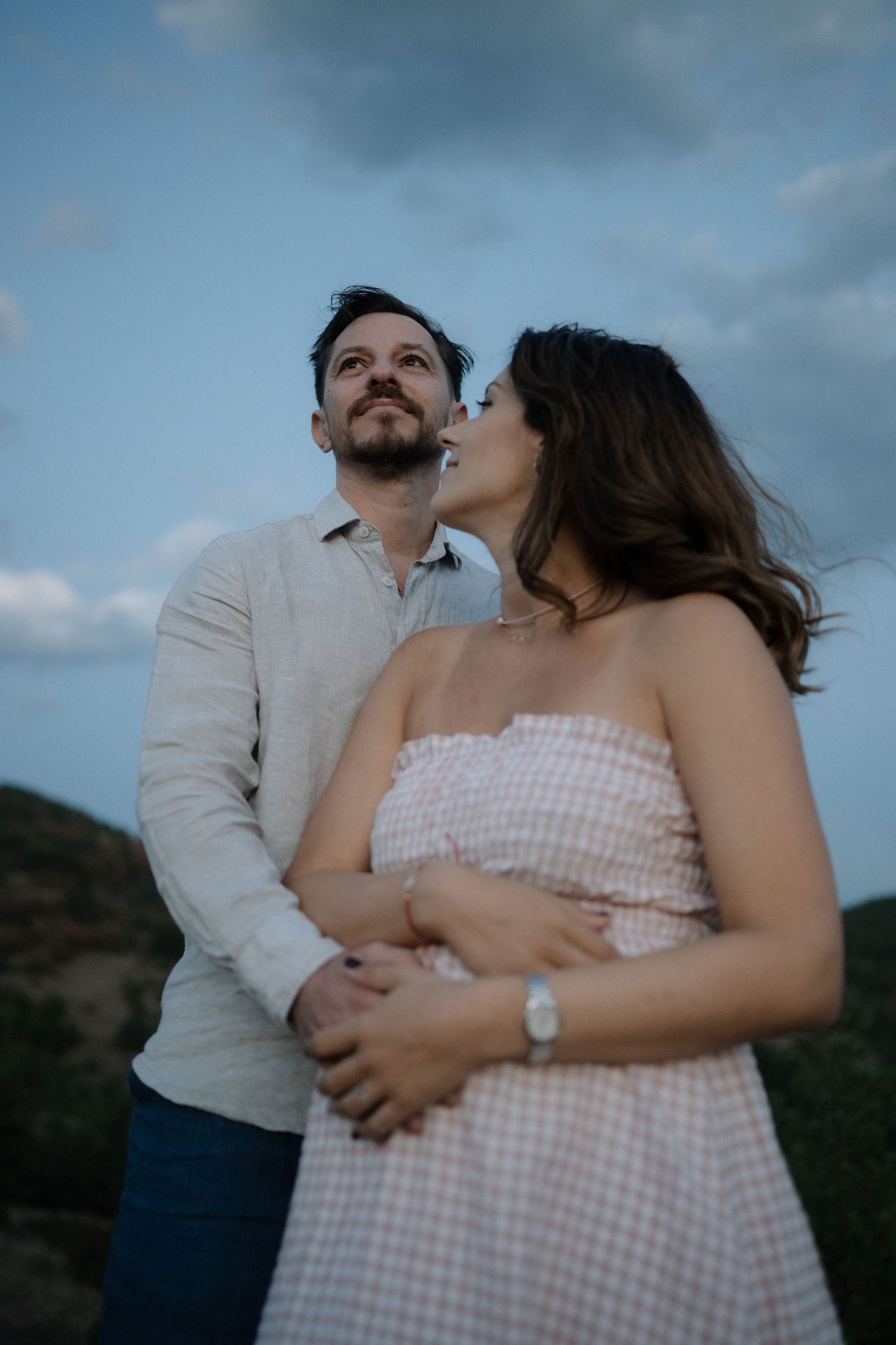 A romantic couple embraces outdoors against a cloudy sky backdrop wearing white and blue casual attire.