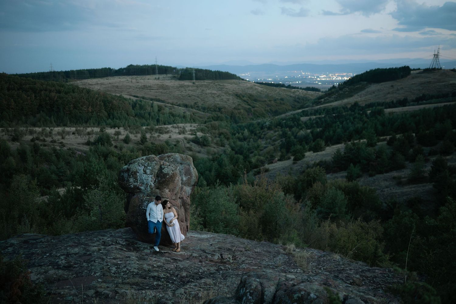 A couple in formal attire poses on a scenic mountain overlook with rolling hills and valleys in the background.