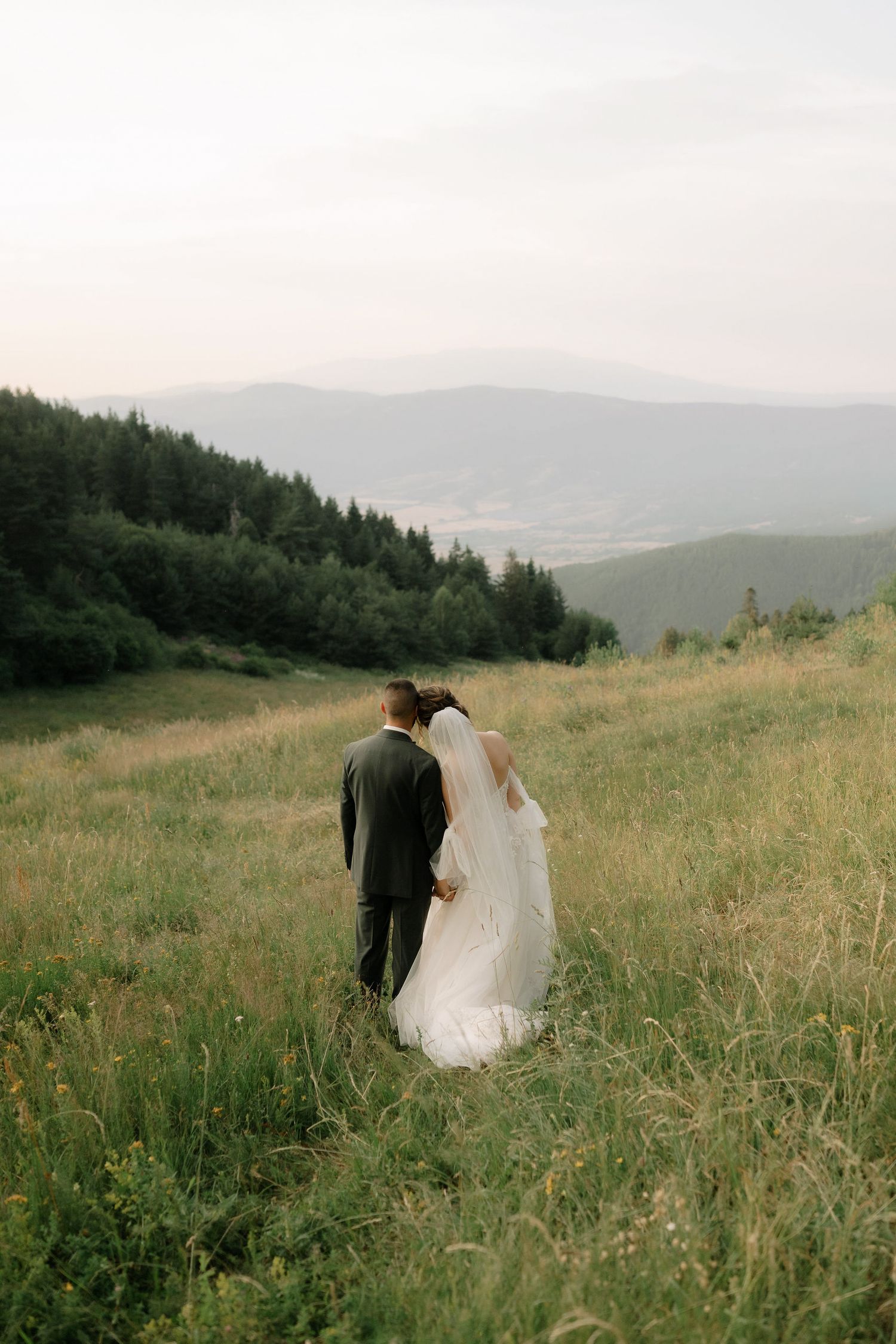 A couple in wedding attire stands on a grassy hillside overlooking misty mountain ranges at sunset.
