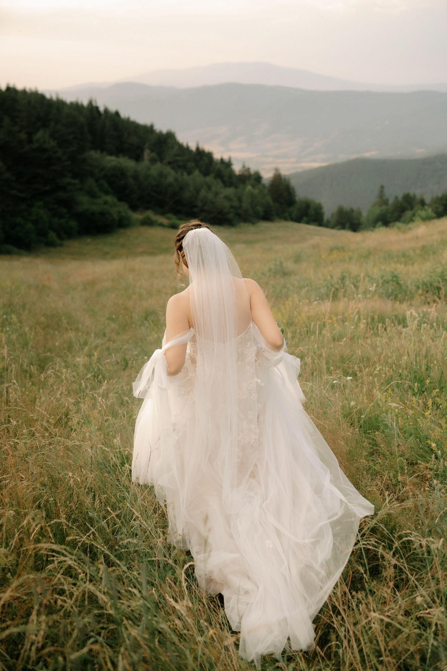 A bride in a flowing white wedding dress walks through a scenic mountain meadow at sunset.