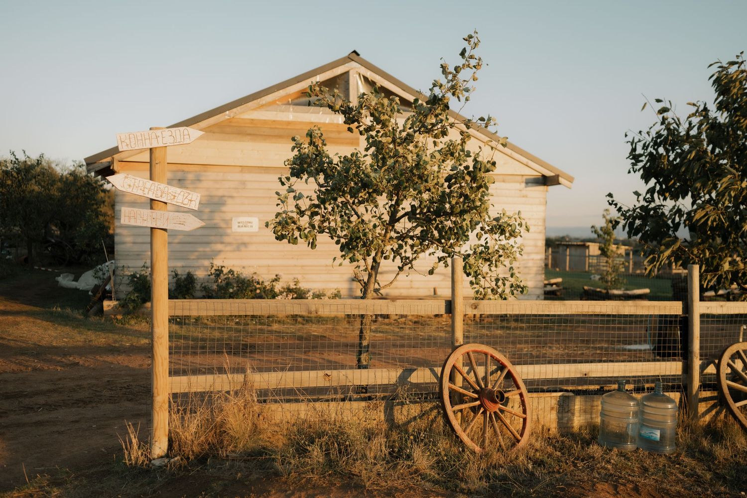 Rustic wooden wagon wheel leans against white farmhouse fence at sunset with trees casting long shadows.