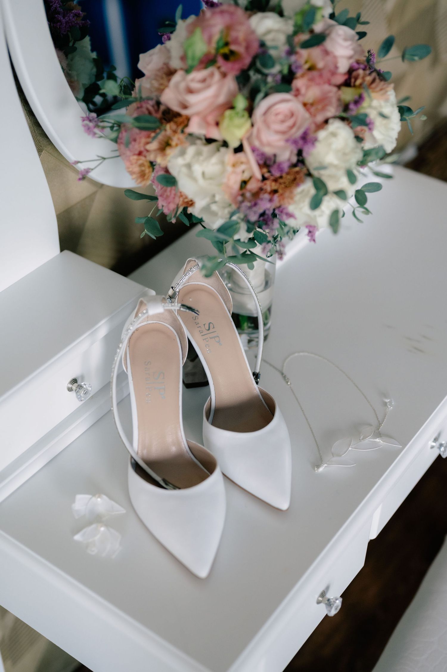 Elegant white bridal heels sit in a drawer beside a soft pastel bouquet of roses and mixed flowers.