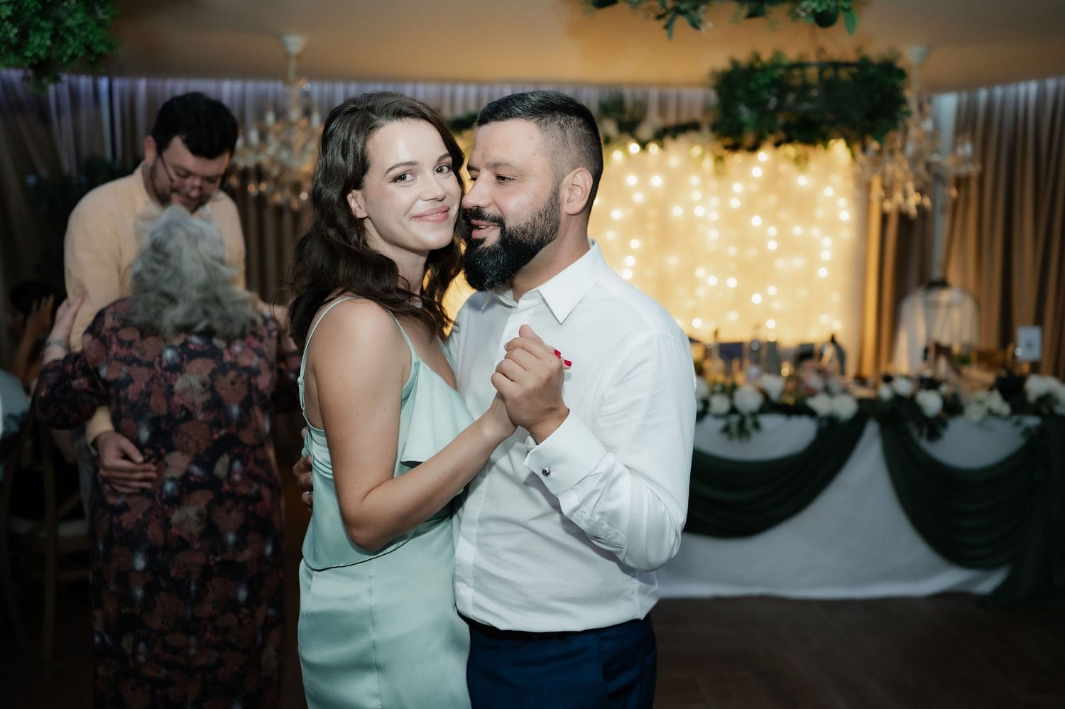 A couple shares a romantic dance moment at a wedding reception with twinkle lights in the background.