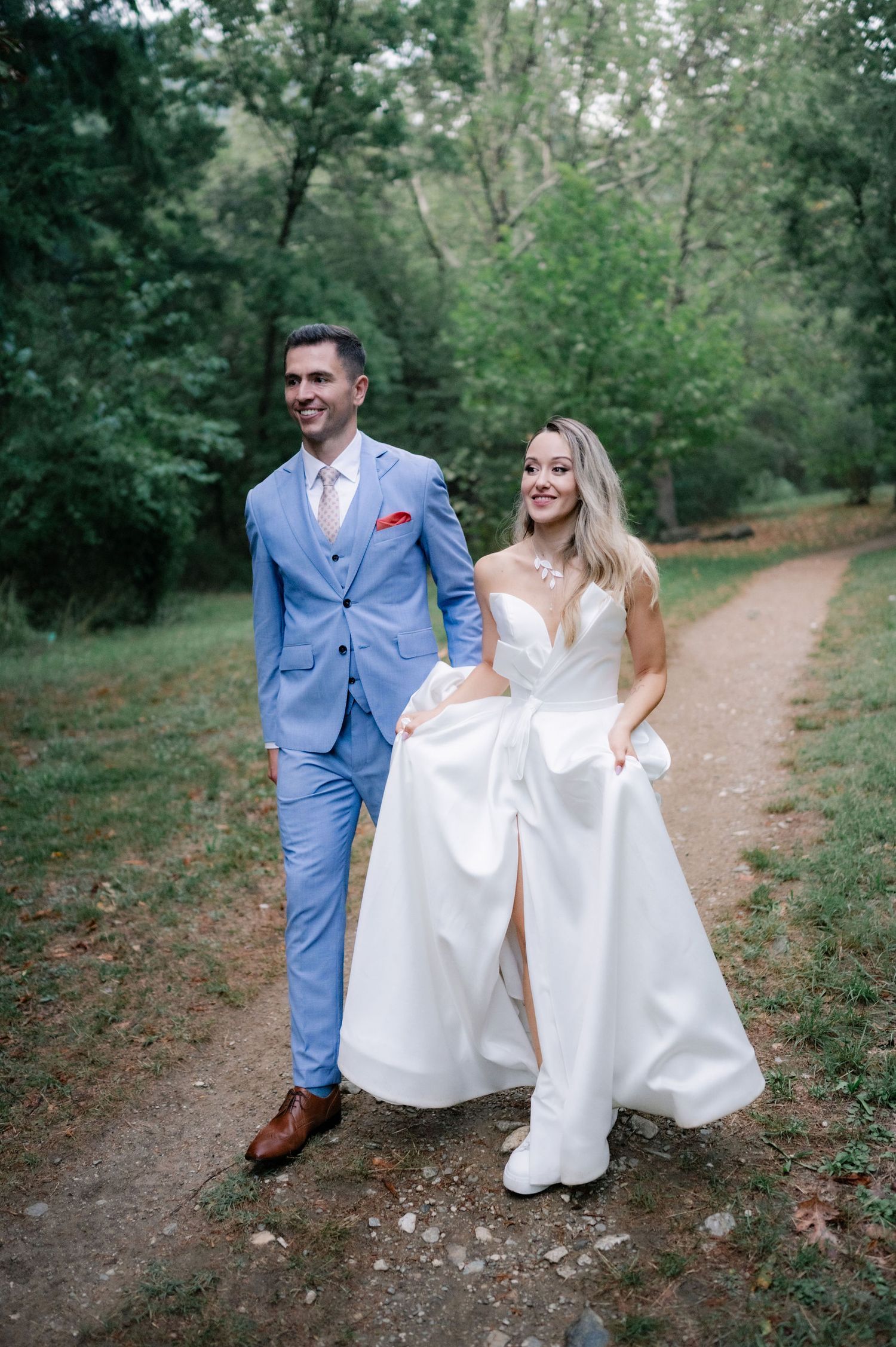 A couple in wedding attire walks together on a wooded path during their outdoor forest photoshoot.