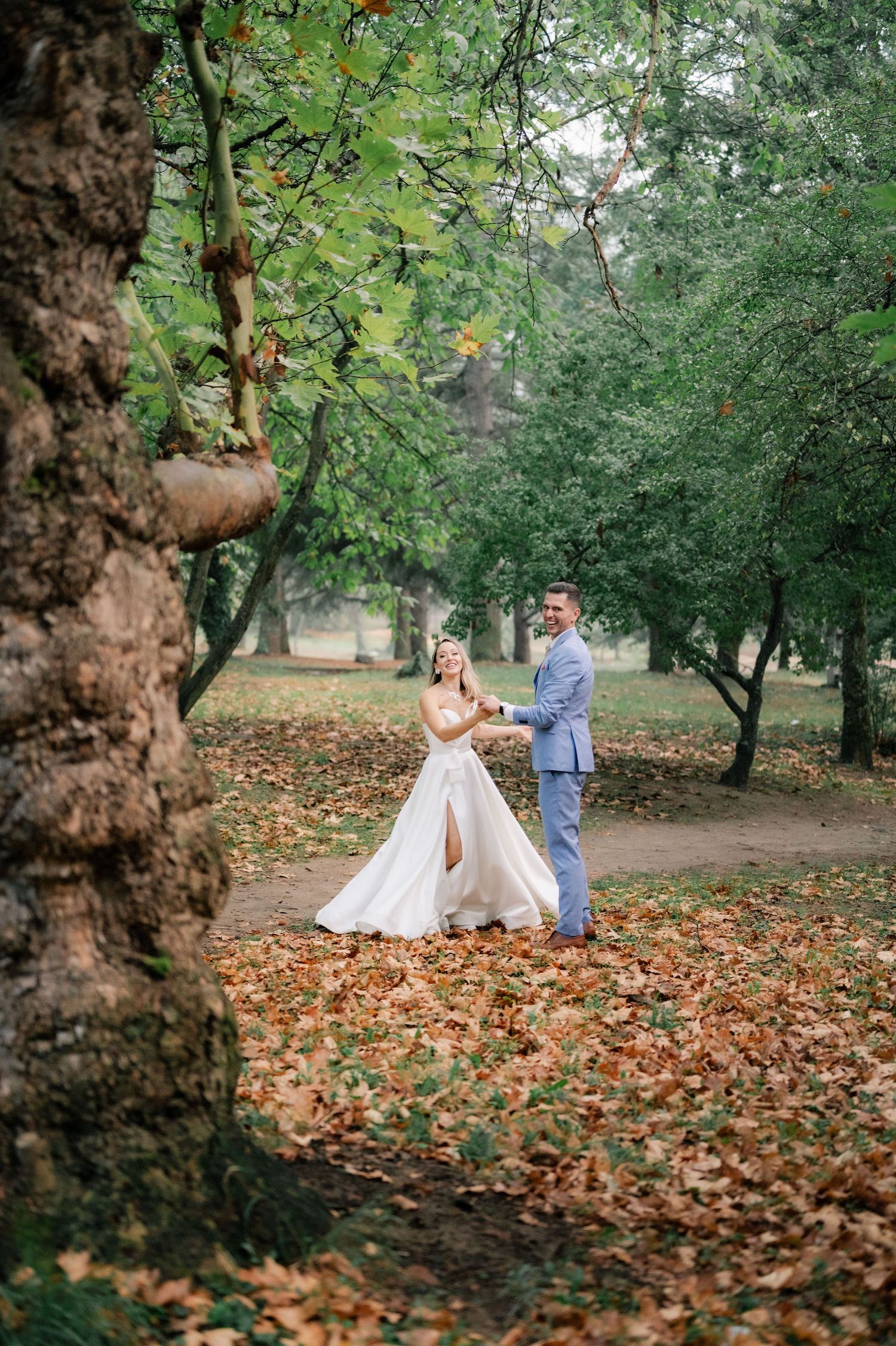 A couple in wedding attire dancing among fallen autumn leaves in a park with tall trees.