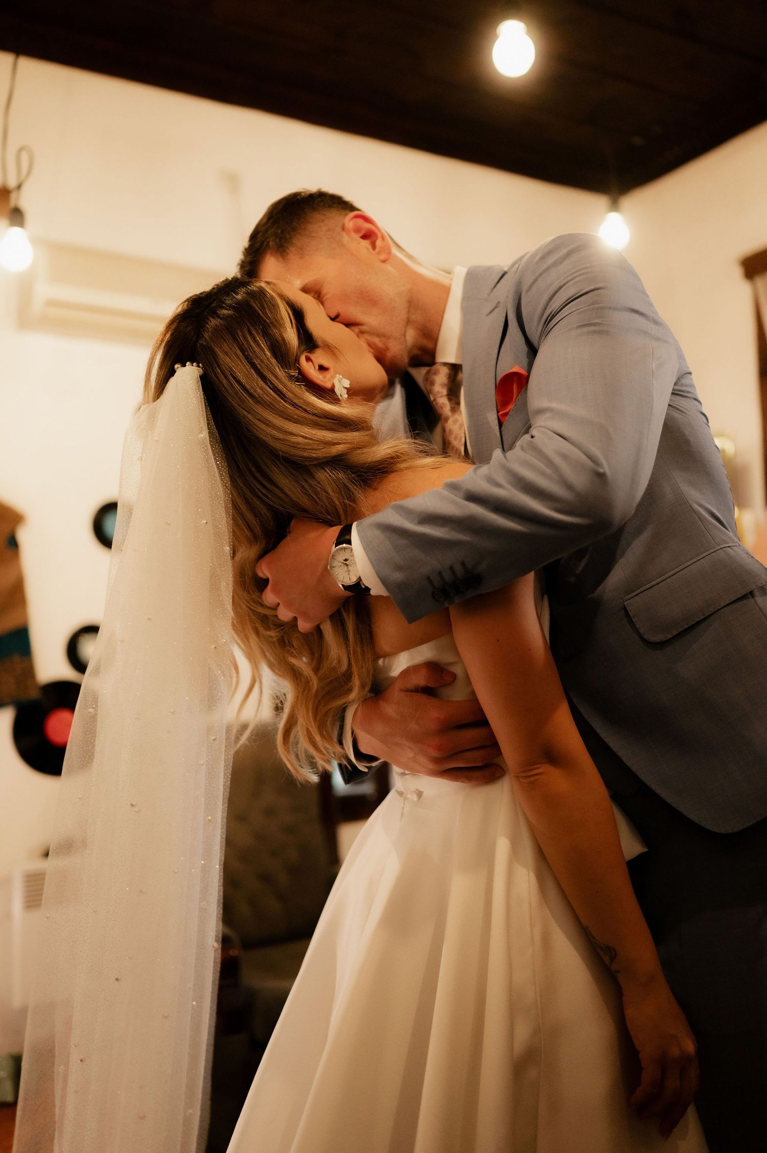 Romantic couple in wedding attire share intimate kisses in a dimly lit room with pendant lights.