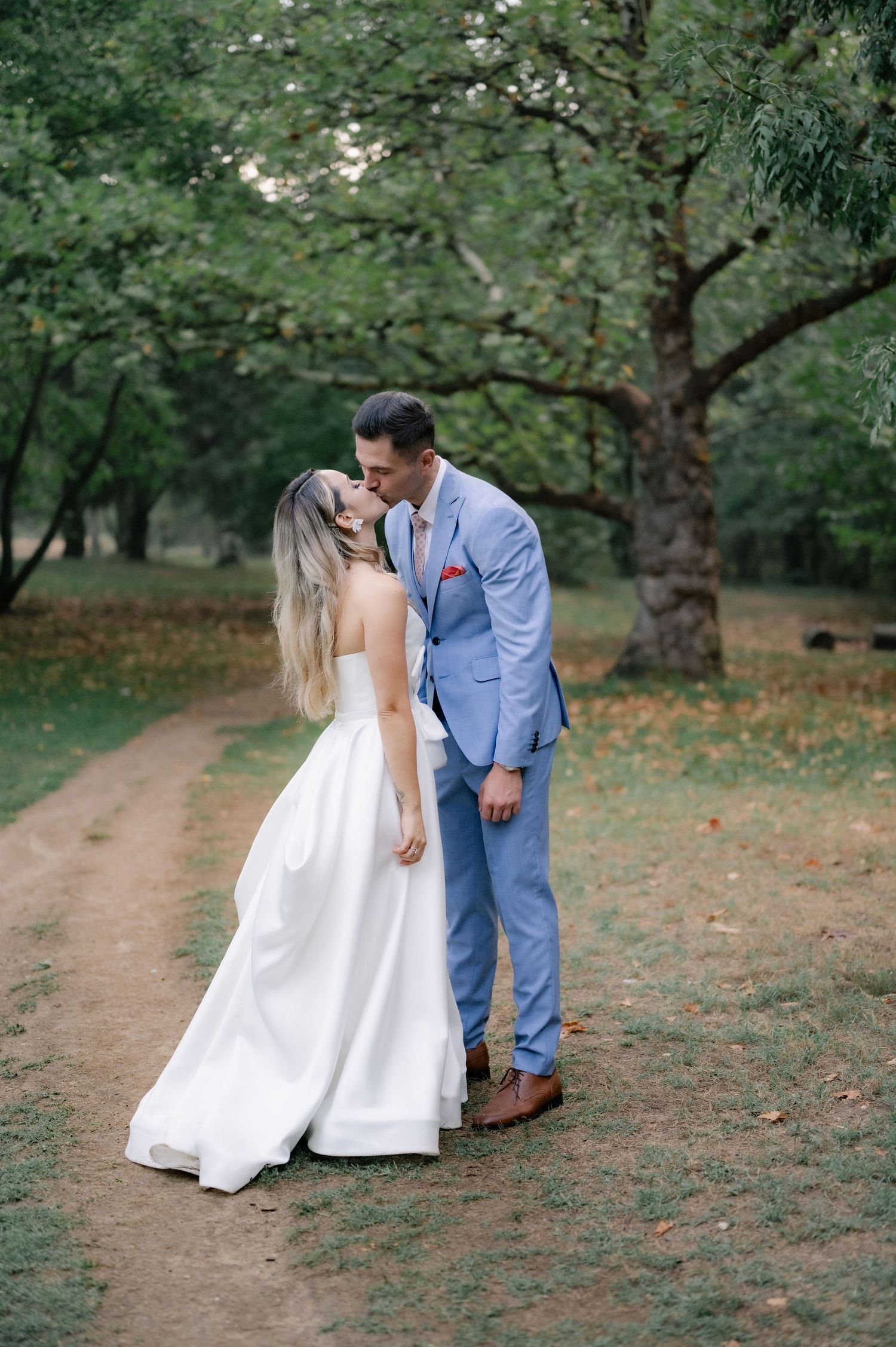 Romantic couple in wedding attire dancing together on a tree-lined dirt path in an outdoor garden setting.