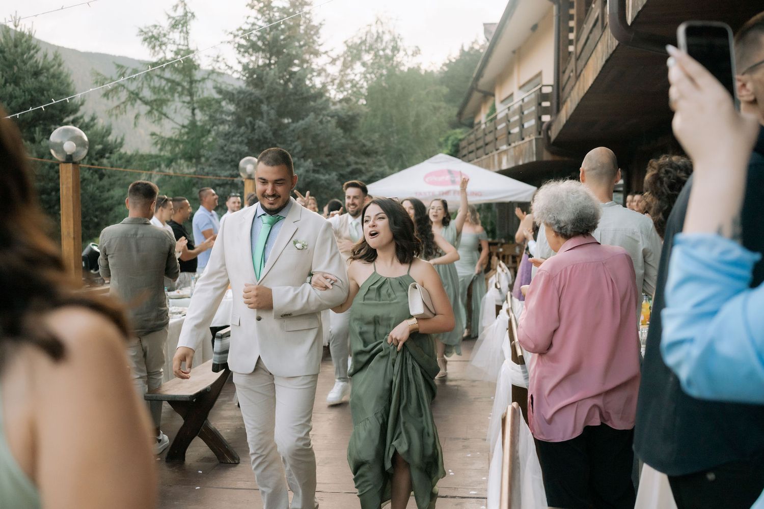 Guests dance and celebrate at an outdoor wedding reception with trees and umbrellas in the background.