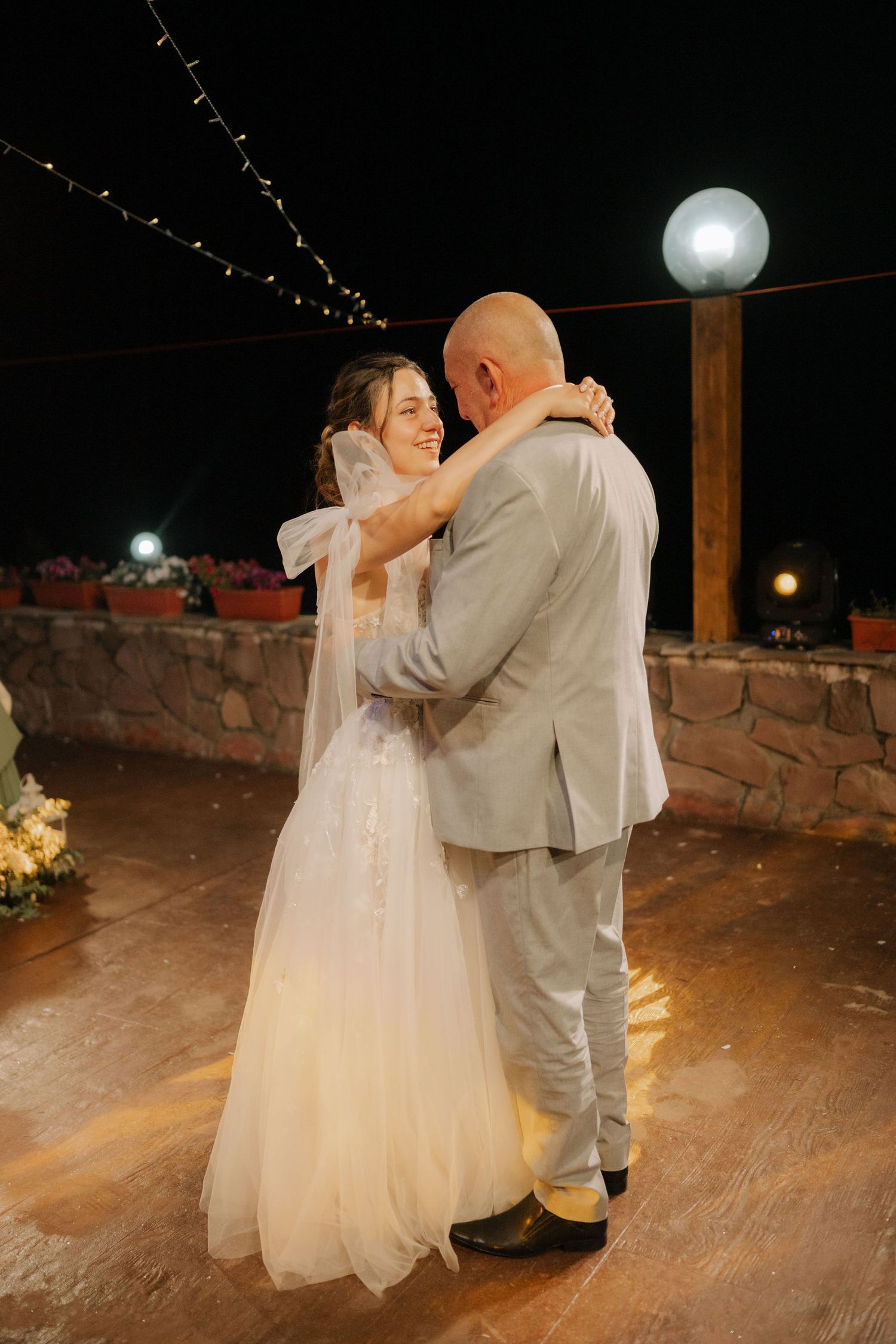 A couple shares their first dance at an outdoor evening wedding reception with string lights and stone walls in the background.