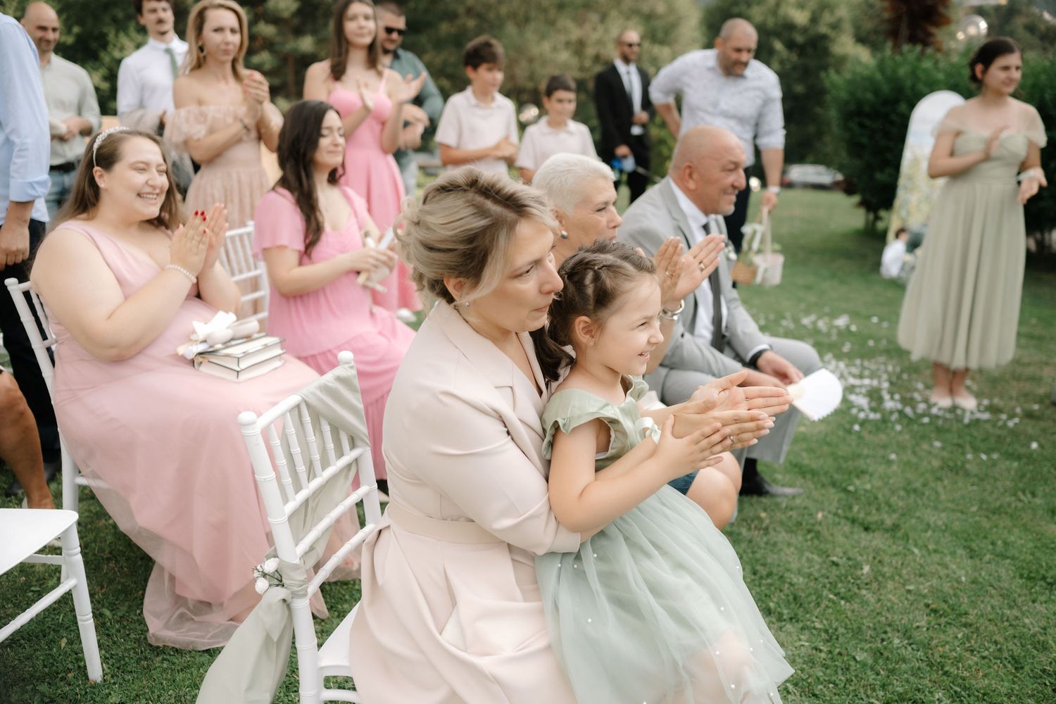 Wedding guests enjoy outdoor reception with pink and beige color scheme while seated in white chairs on grass lawn.