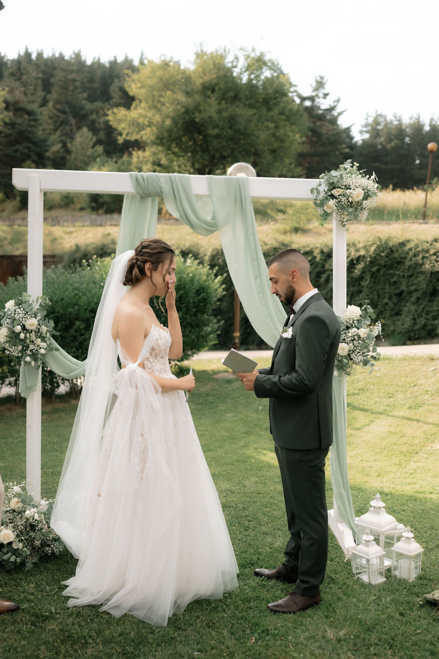 A romantic outdoor wedding ceremony features a white draped arch with floral arrangements in a garden setting.