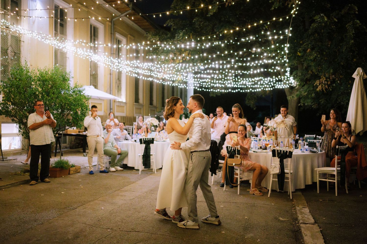 Romantic outdoor evening wedding reception with string lights overhead as couple dances on patio surrounded by guests.