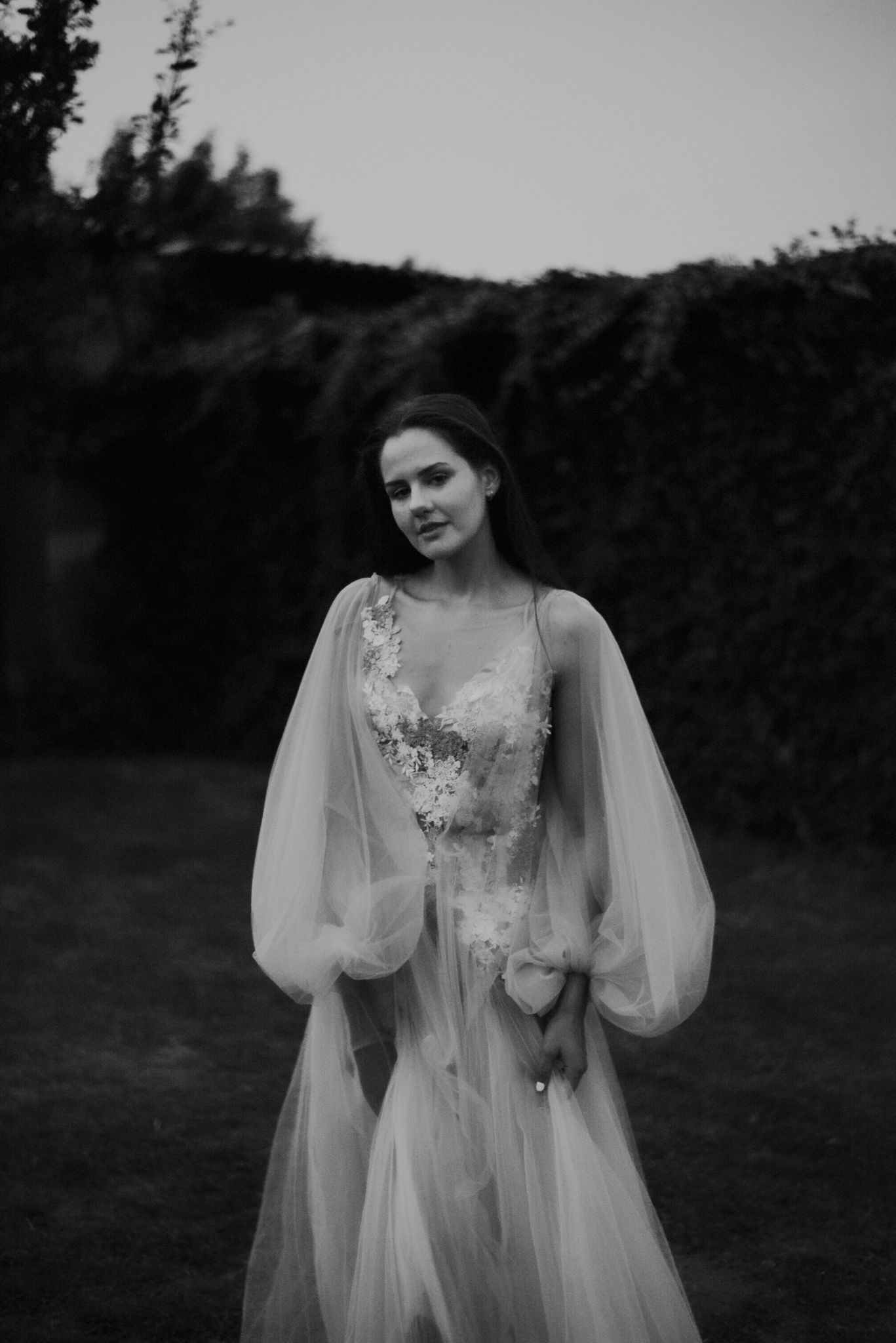 Ethereal black and white portrait of a bride in a romantic lace gown with billowing sheer sleeves in a moody outdoor setting.