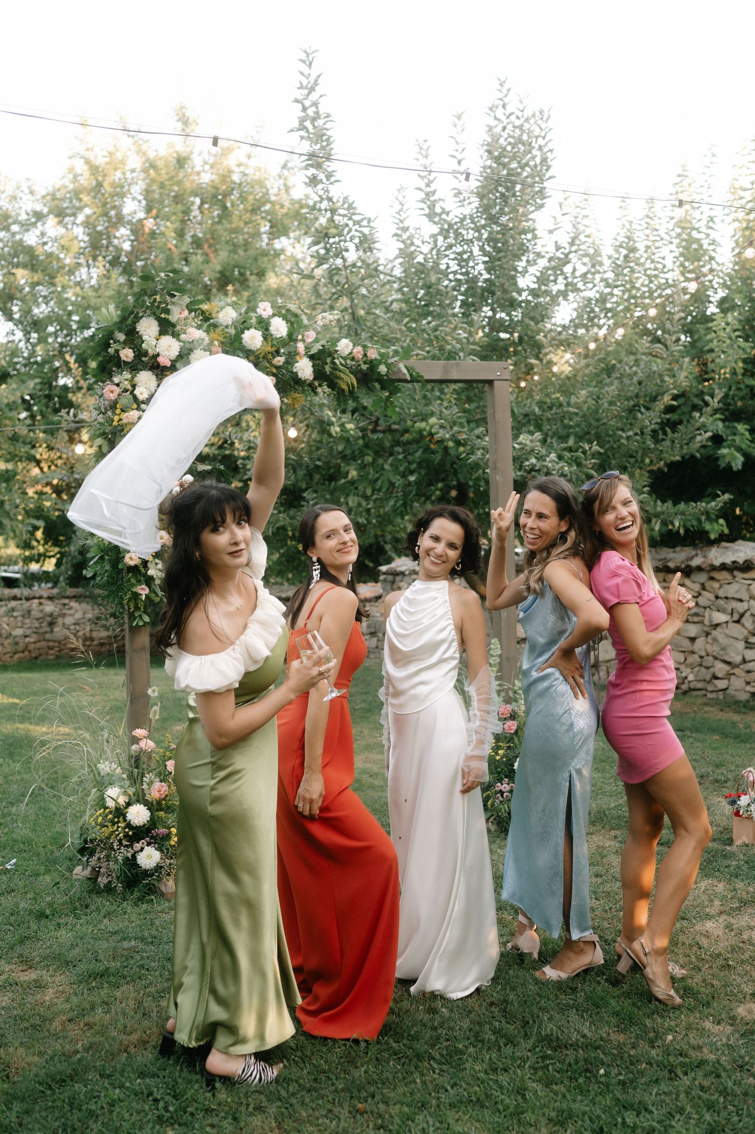 A group of bridesmaids in colorful dresses pose playfully together on a lawn with floral decorations in the background.
