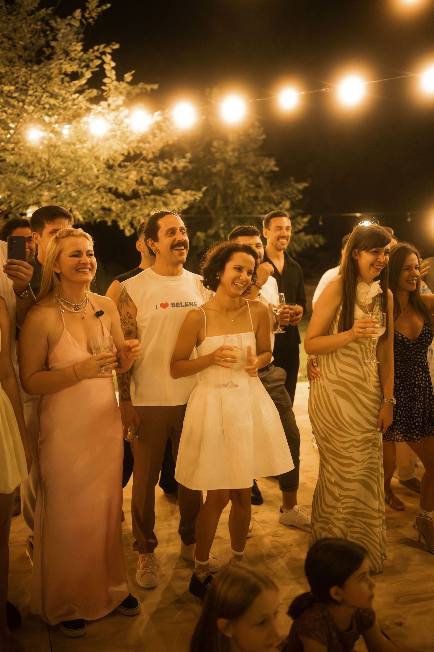People dancing and celebrating at a nighttime outdoor party under warm string lights and glowing lanterns.
