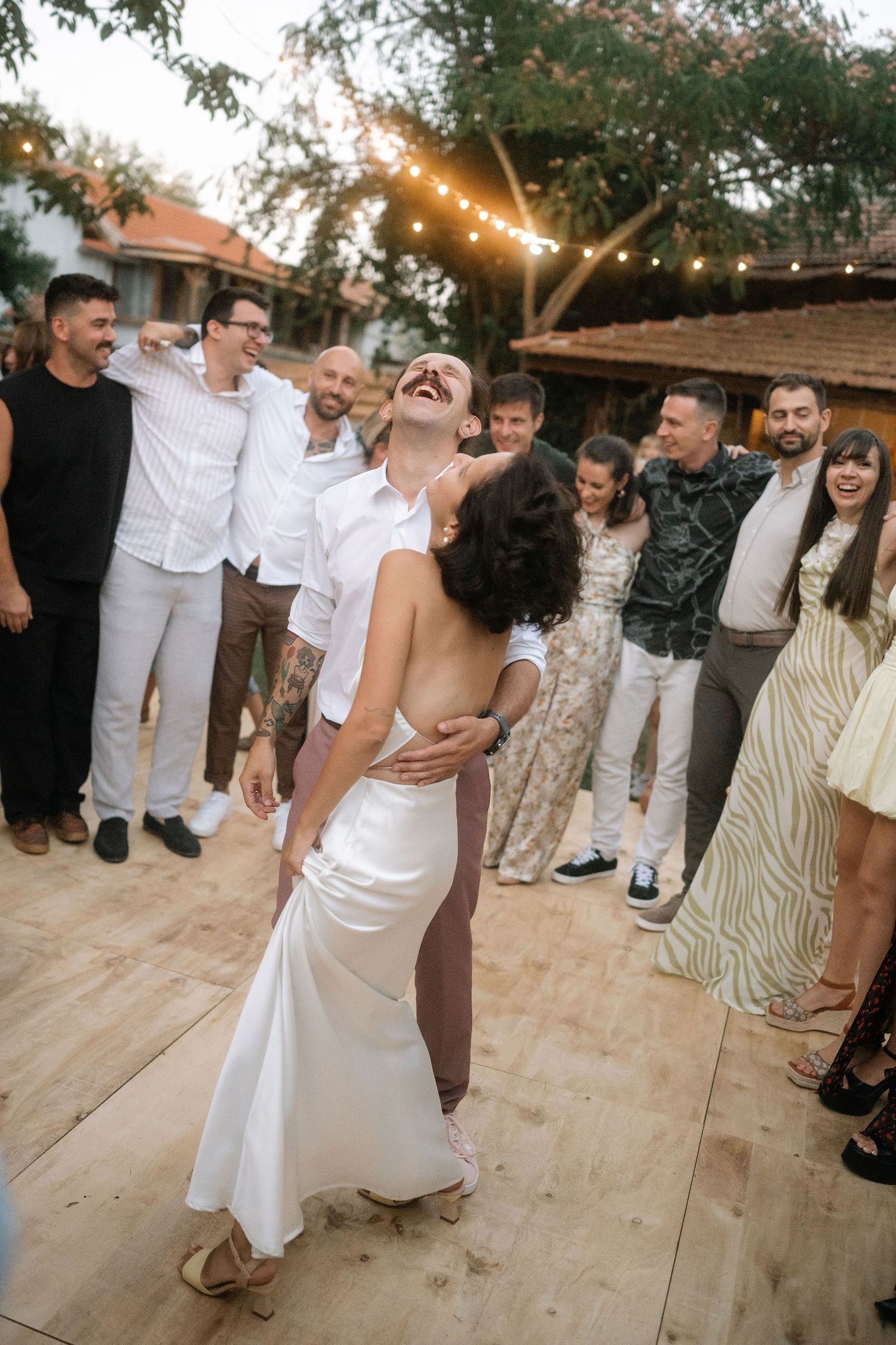 Group of wedding guests dancing and laughing together on wooden dance floor under string lights at outdoor reception.