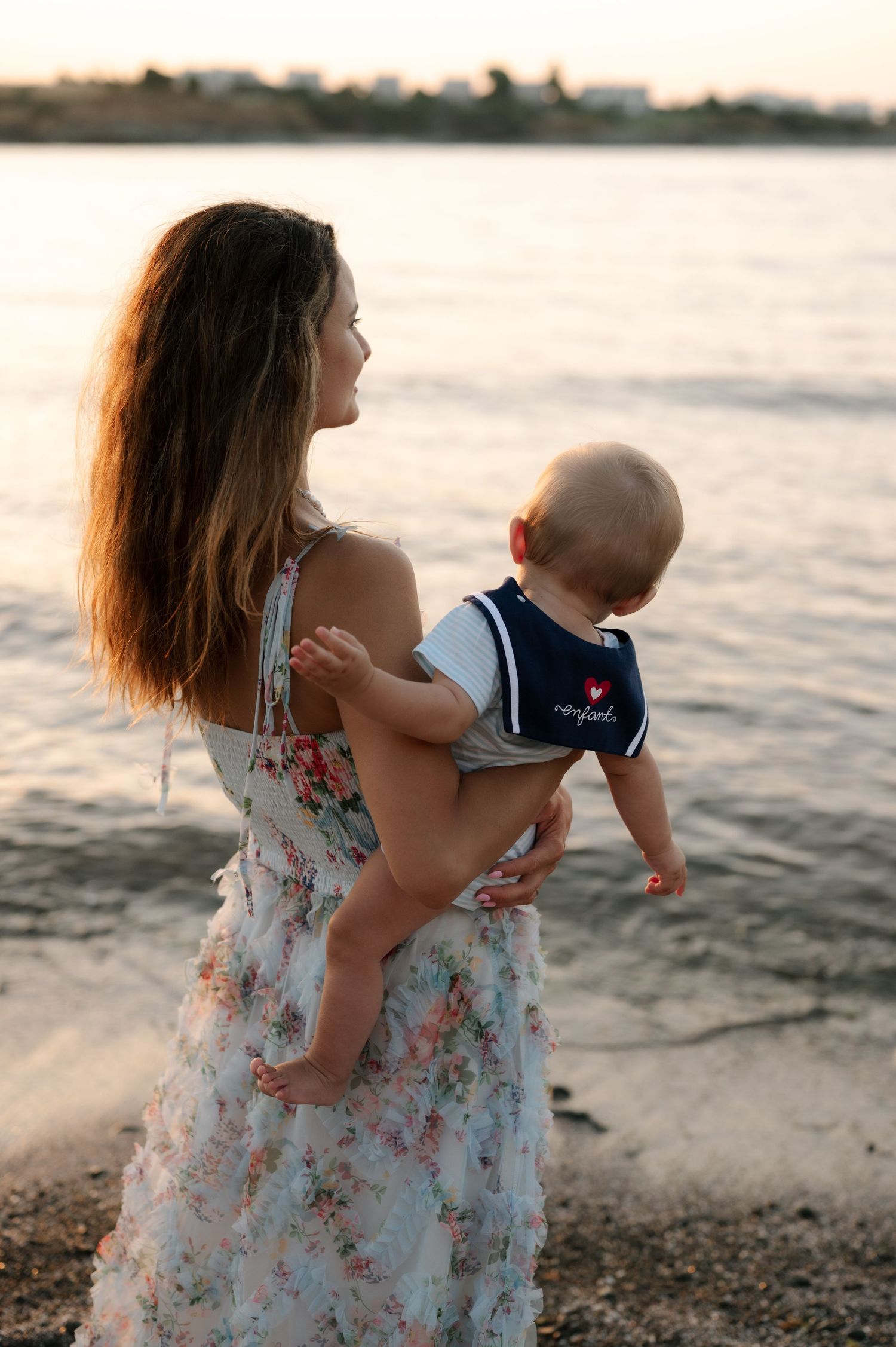 Mother and child in floral dresses share tender moments on a beach at sunset.