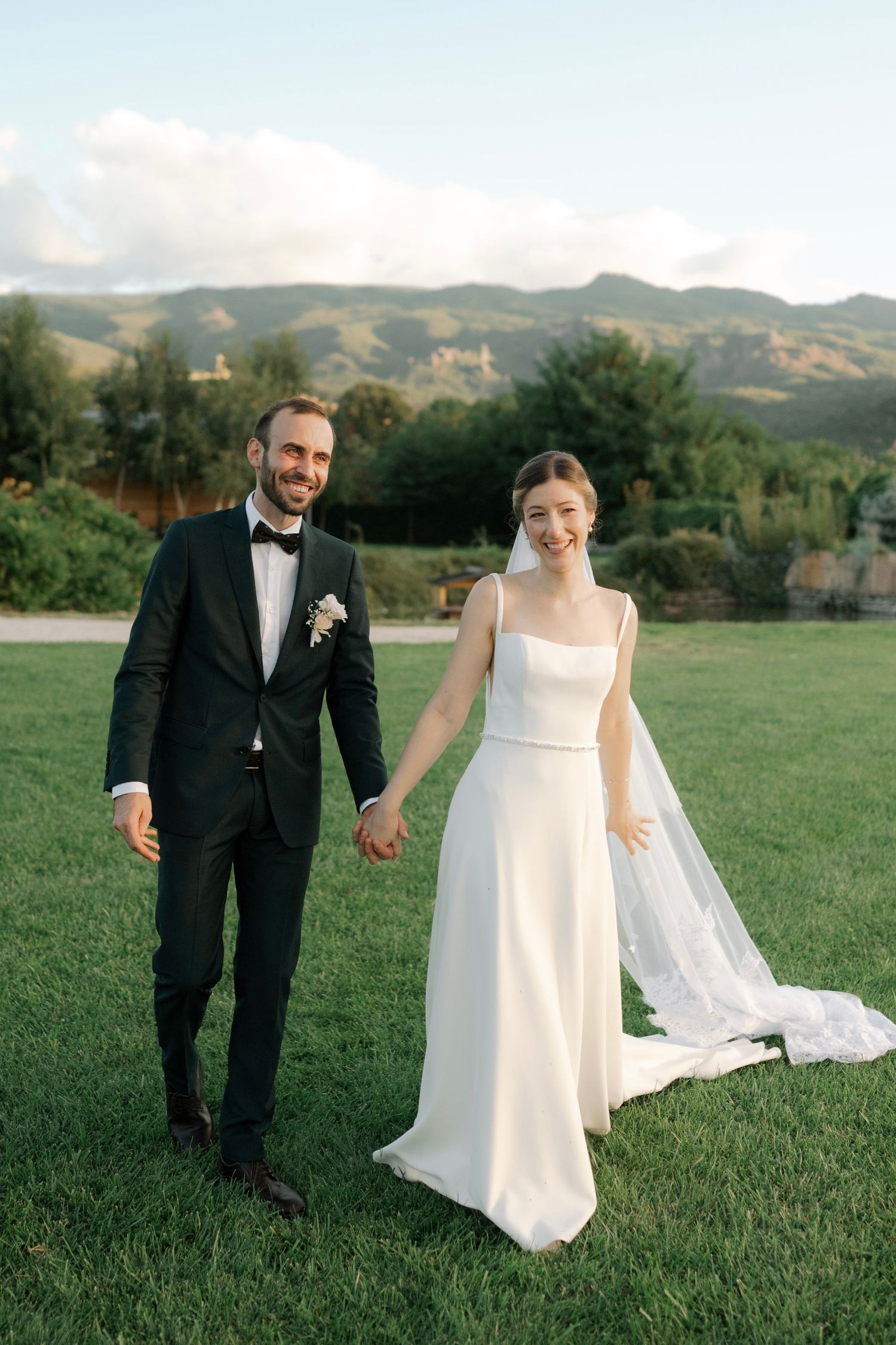 A couple walks hand in hand across a green lawn with mountains in the background on their wedding day.