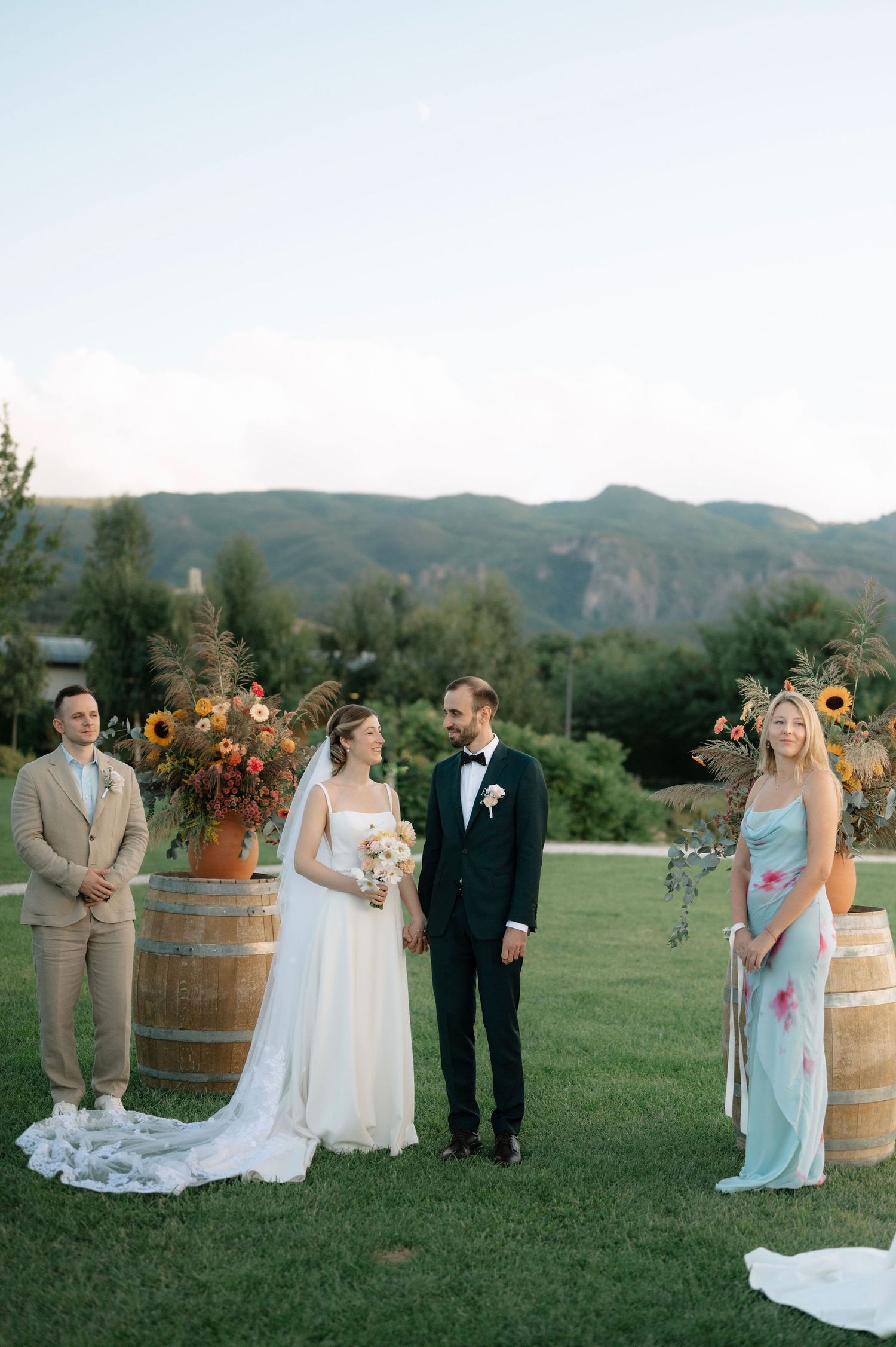 Wedding party poses outdoors in front of wine barrels and sunflowers on a scenic mountainside venue.
