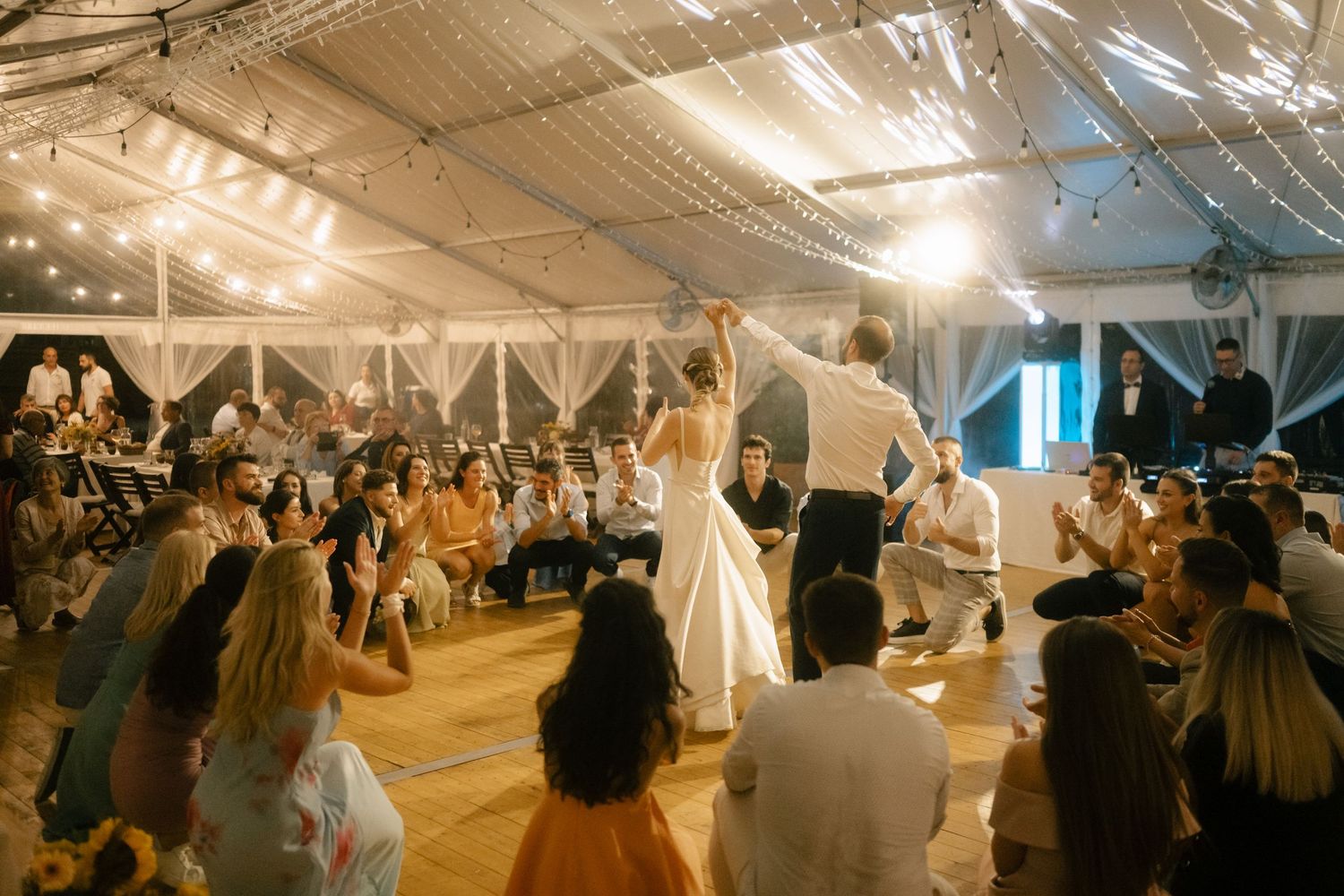 Large white wedding reception tent filled with guests dining at long tables under glowing string lights.