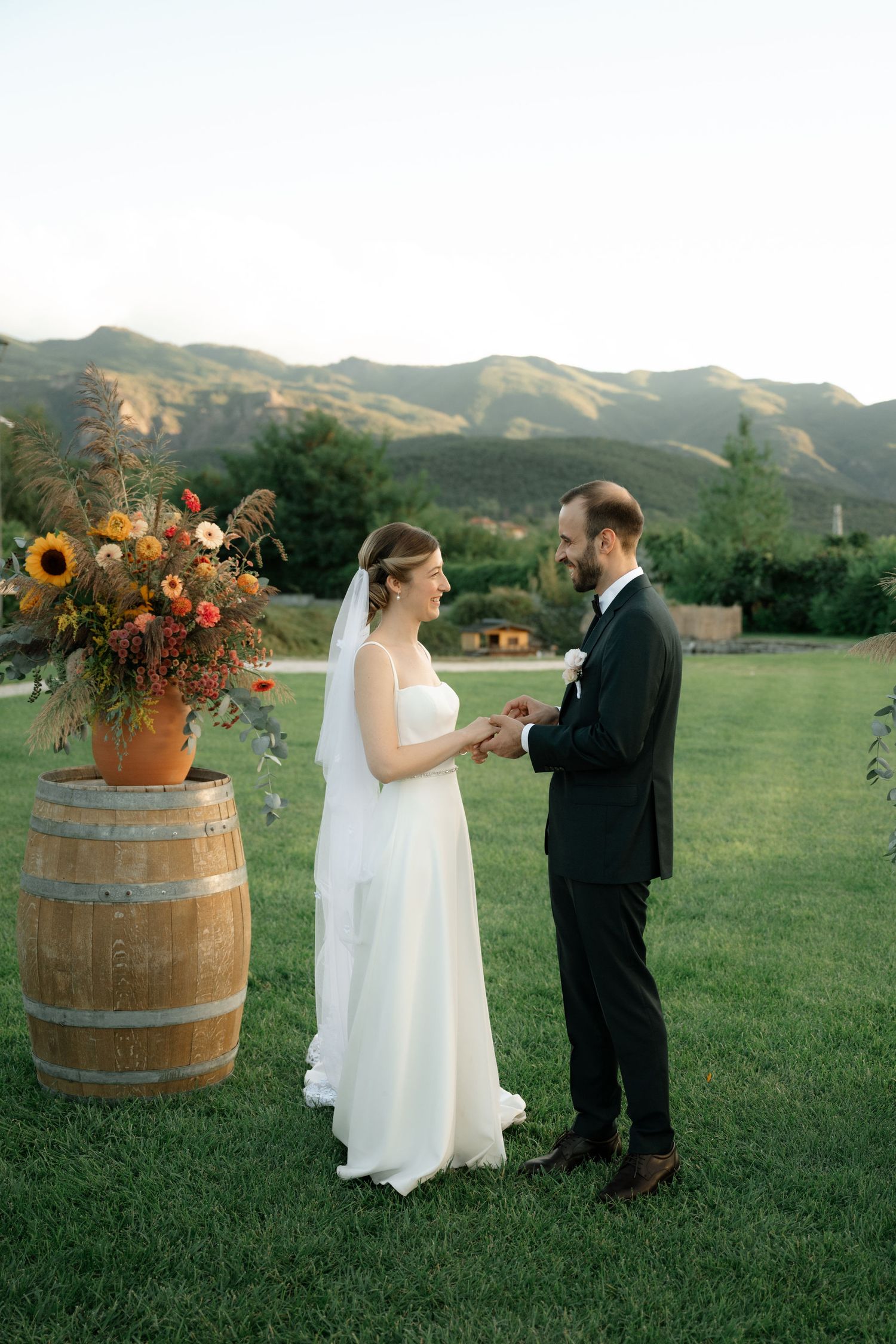 A wedding couple exchanges vows in a scenic mountain setting with a rustic wine barrel and sunflower arrangement.
