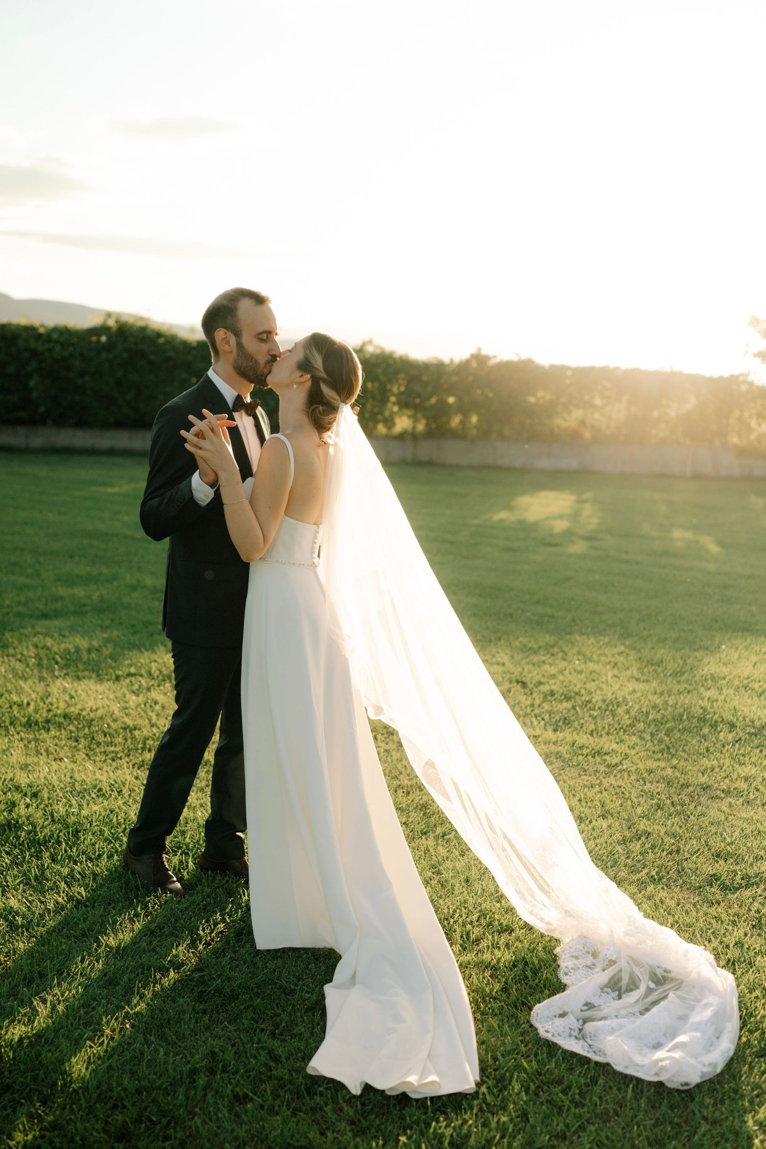 A romantic wedding couple embraces during golden hour sunset in a grassy field with a flowing white veil.