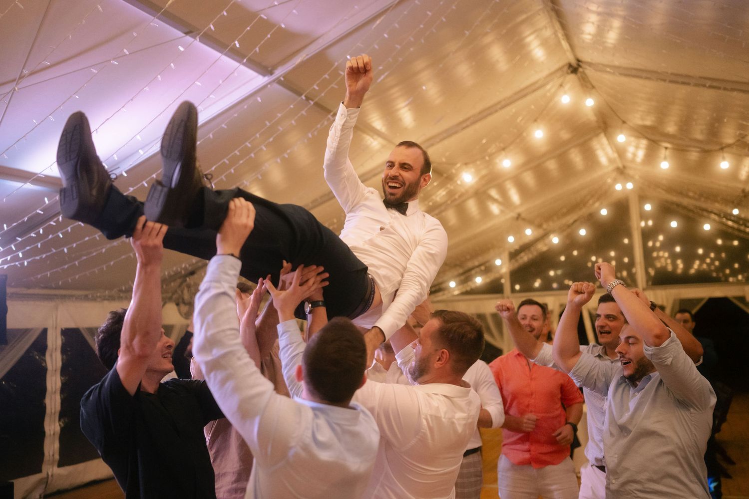Wedding guests dancing and celebrating under a white tent with string lights creating a festive atmosphere.