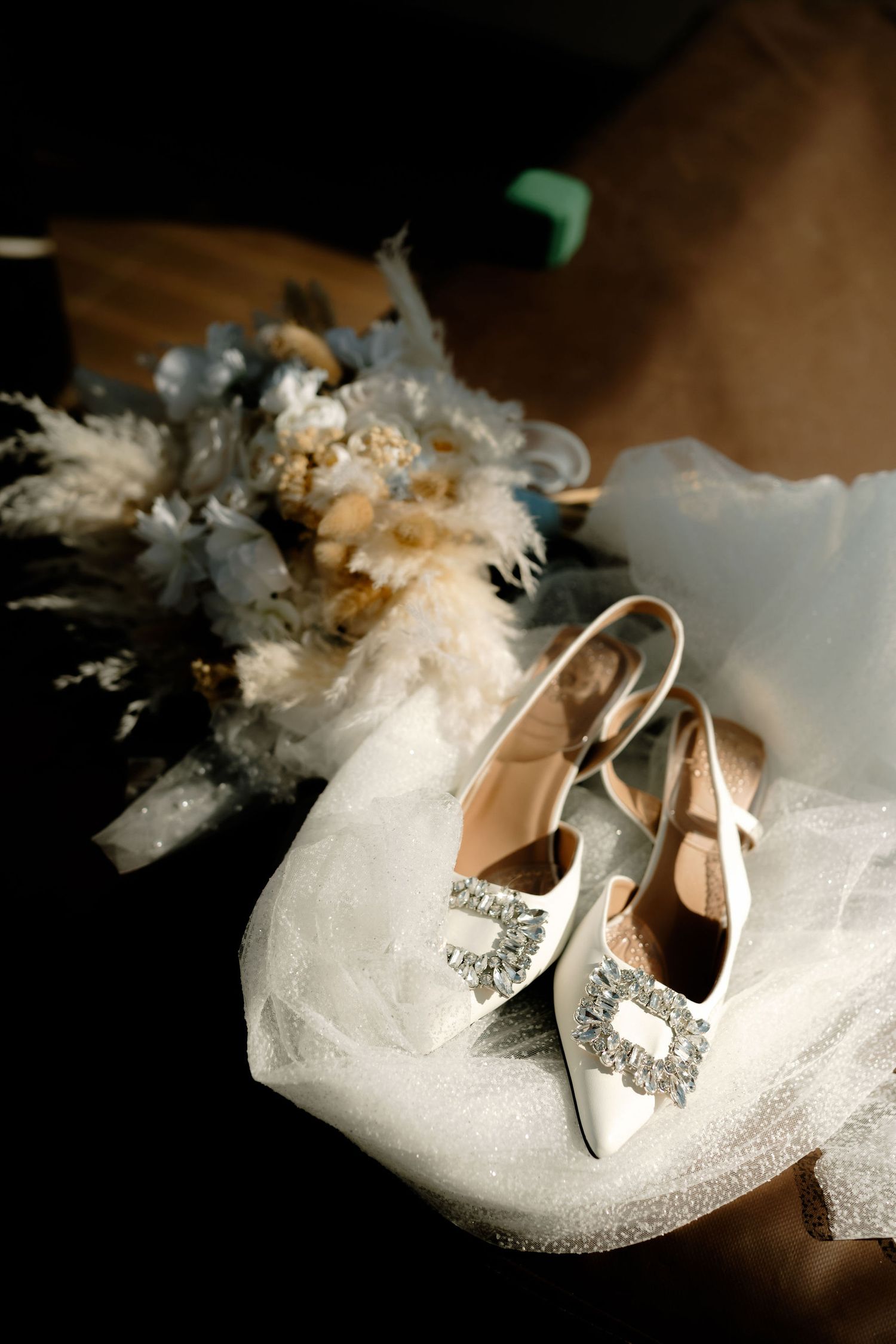 Elegant white bridal shoes with crystal embellishments displayed with delicate tulle fabric in dramatic lighting.