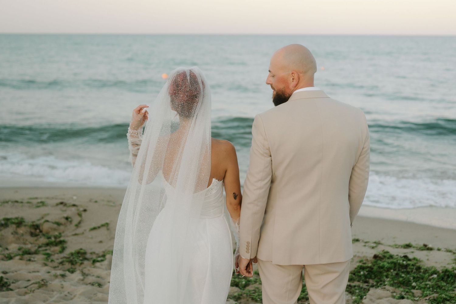A couple in light-colored attire walks along a serene beach at sunset with ocean waves rolling in the background.