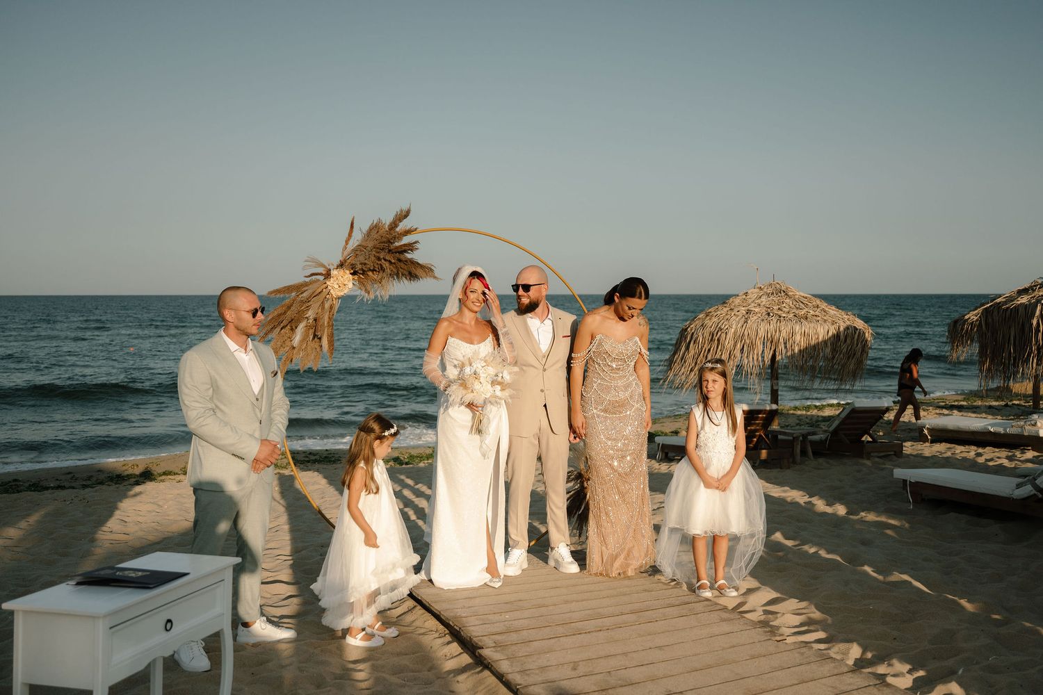 A beachside celebration at sunset with people gathered on a wooden deck near thatched umbrellas by the ocean.