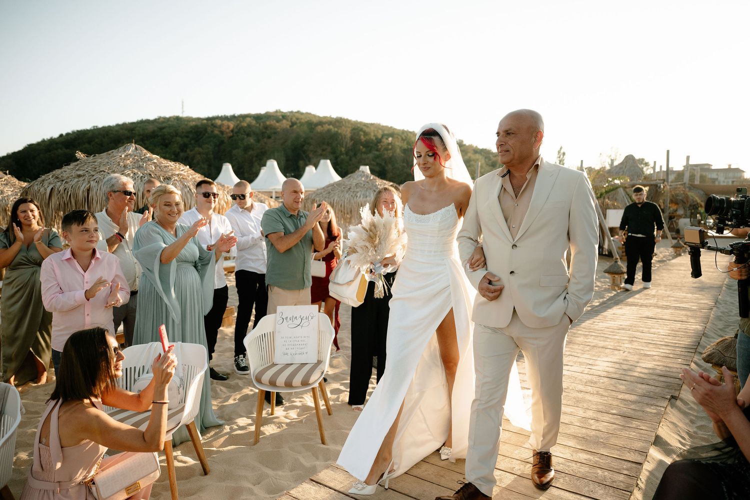 Wedding guests celebrate as a couple walks down the aisle at an outdoor ceremony with white tents in the background.