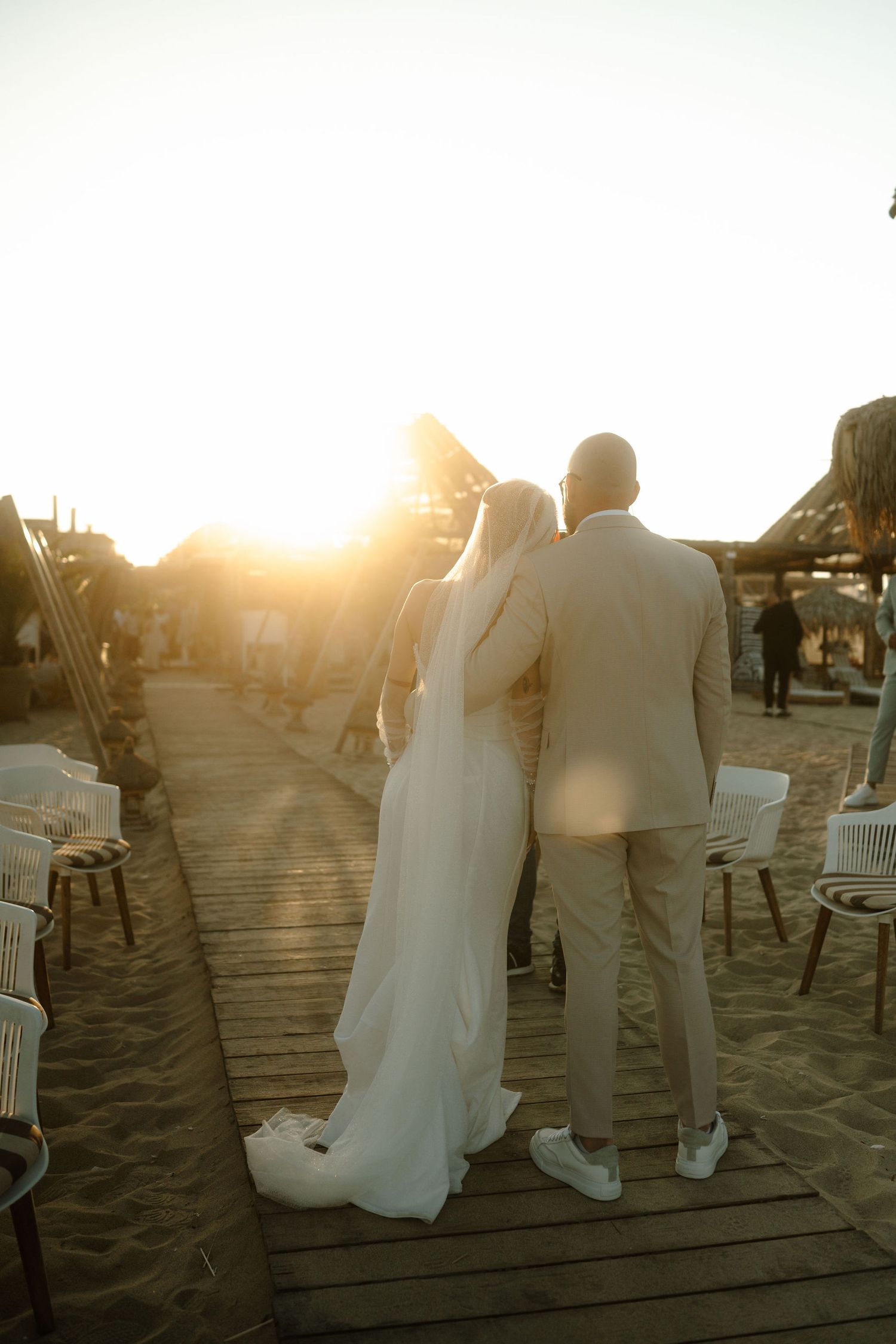 A couple in wedding attire stands on a wooden boardwalk at sunset creating silhouettes against the golden sky.